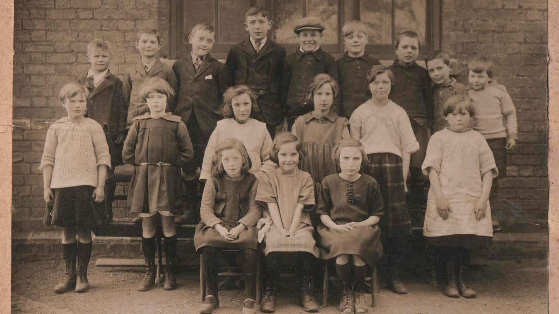 Black and white sepia image showing a school photograph, showing three rows of young children in period dress