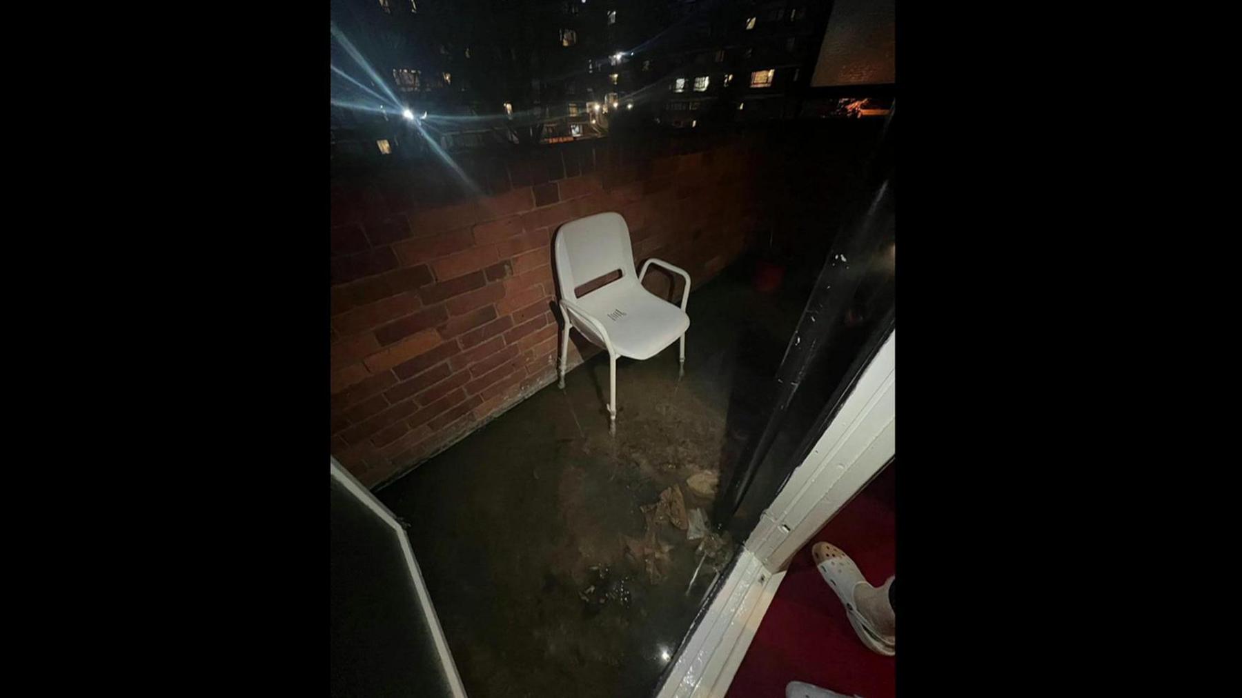 Raw sewage on a balcony. An outdoor chair is against a brick wall, the bottom of its legs are submerged in murky, dirty water.