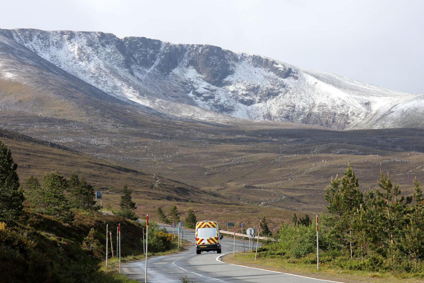 Snow dusts a coire, a bowl-like hollow in a mountainside, in the Cairngorms. On a road below the mountain is a van with brightly-coloured rear doors.