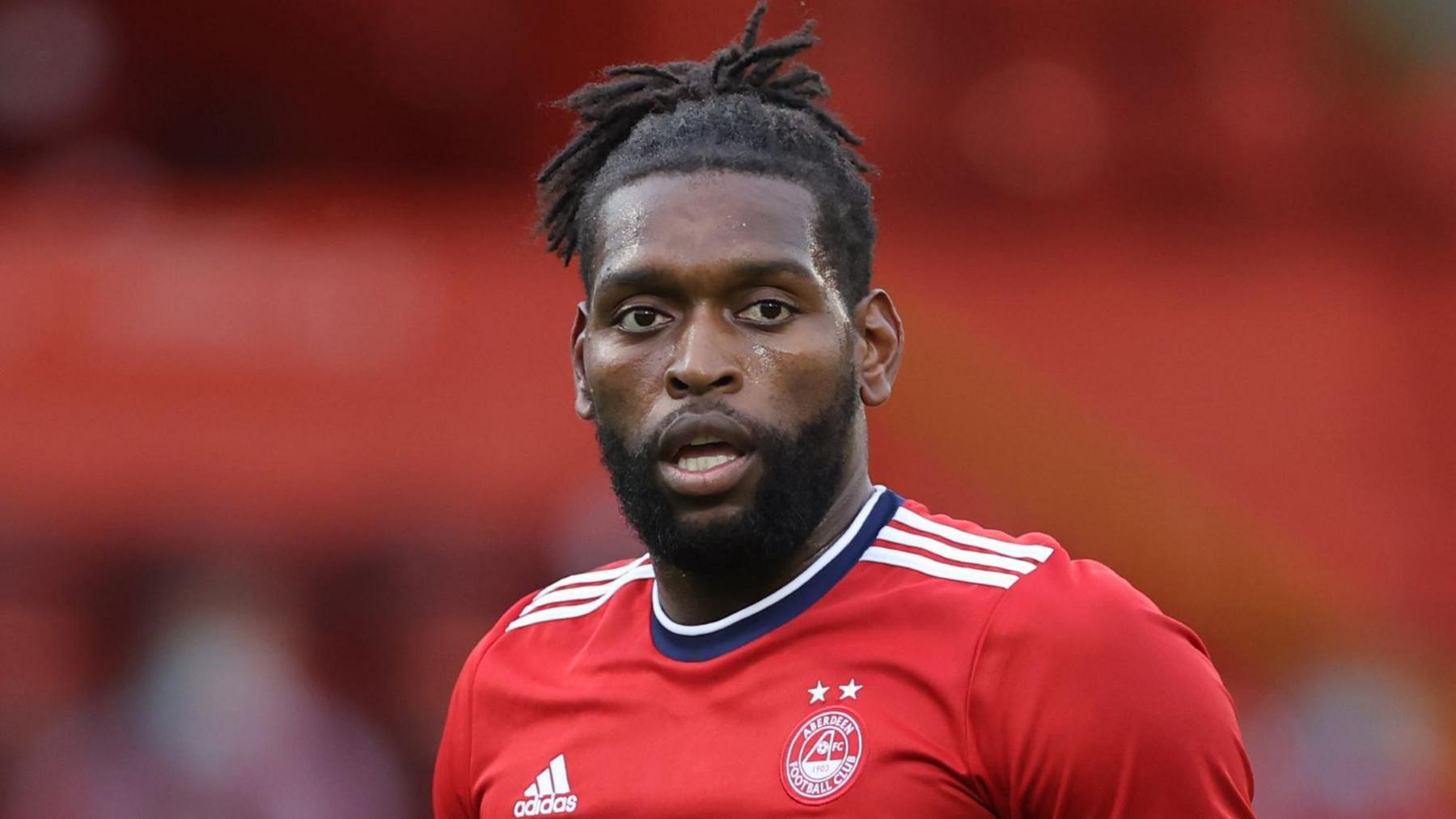 Jay Emmanuel-Thomas wearing a red Aberdeen shirt while playing a match. He is looking across the pitch with his mouth open.