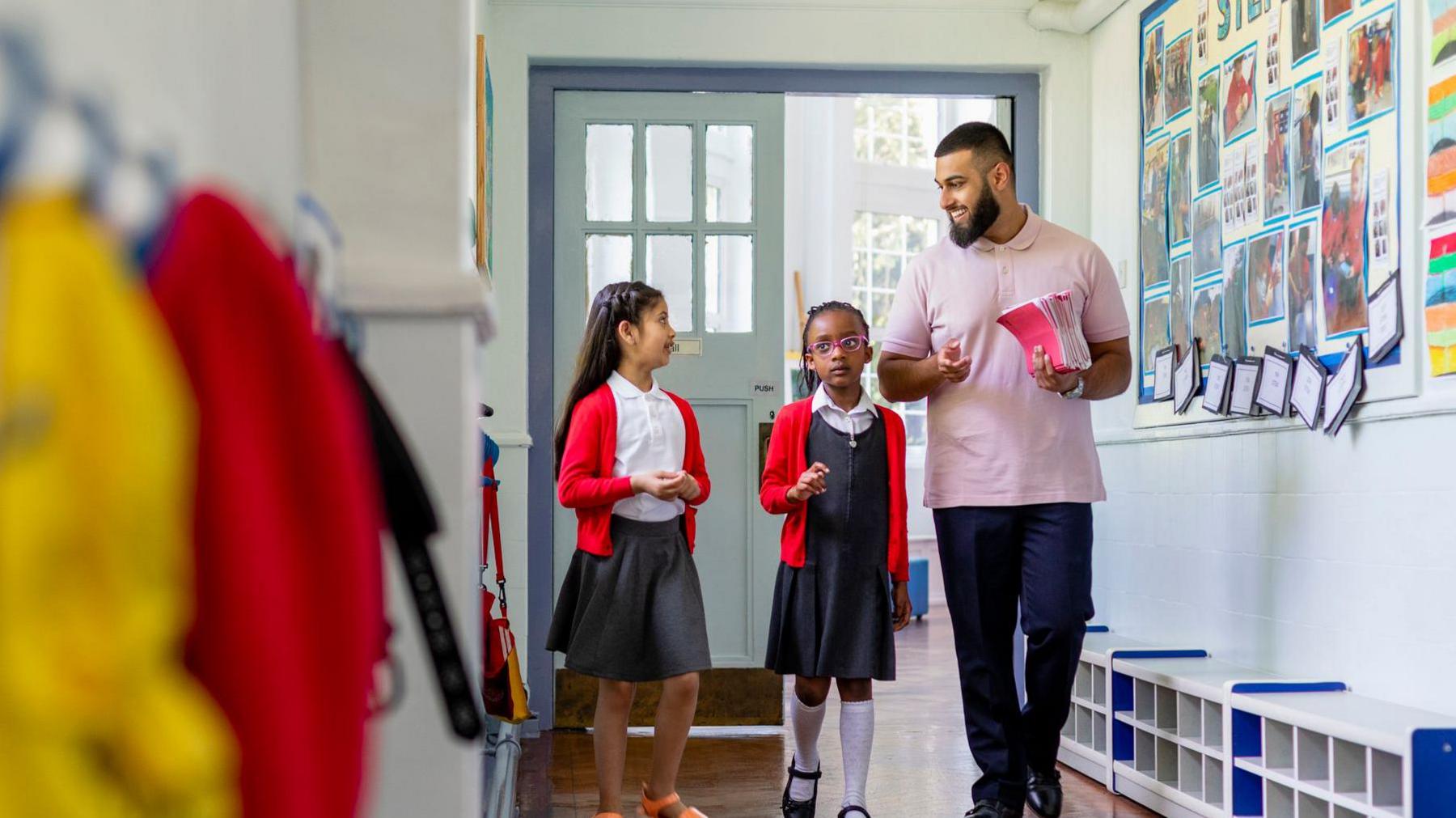 A stock image of two female primary school pupils, both in red cardigans, walking along a school corridor with their male teacher who is wearing a pink polo shirt and carrying a stack of pink notebooks.