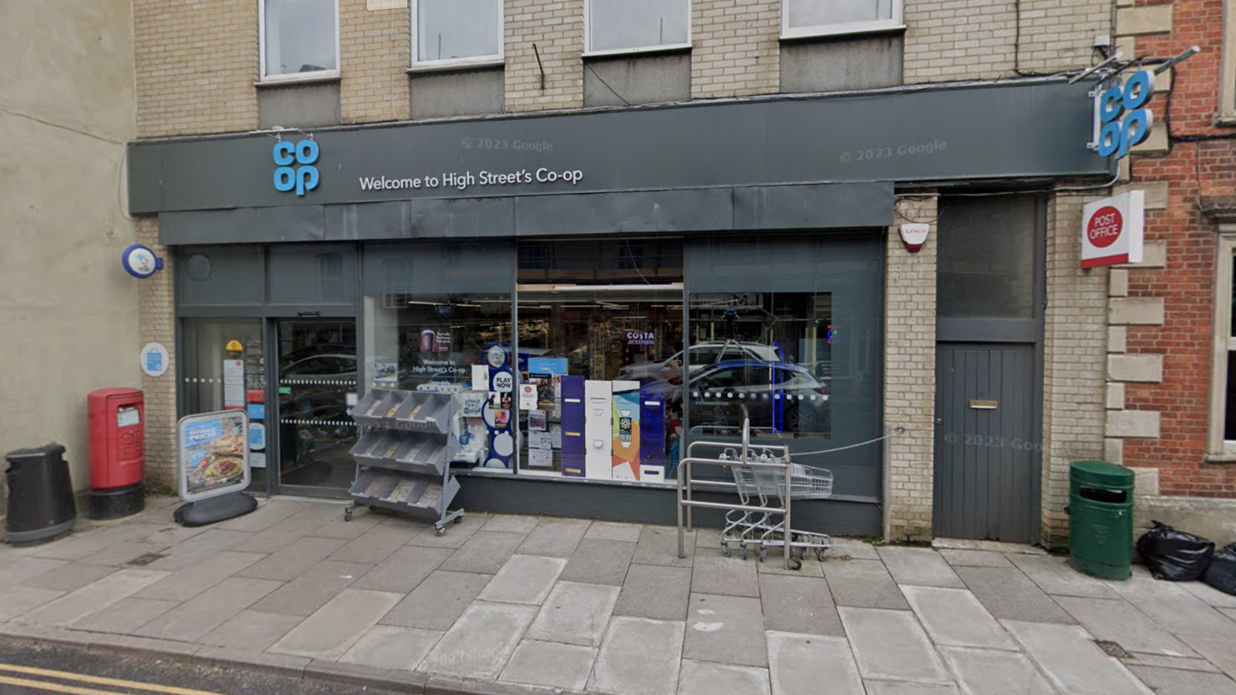 Google maps street view of the Co-op along Malmesbury's High Street. The building is grey with a blue Co-op logo on the top. A trolley, a red post box a newspaper stand are outside the shop. A Co-op sign and post office sign are on the side of the building. 