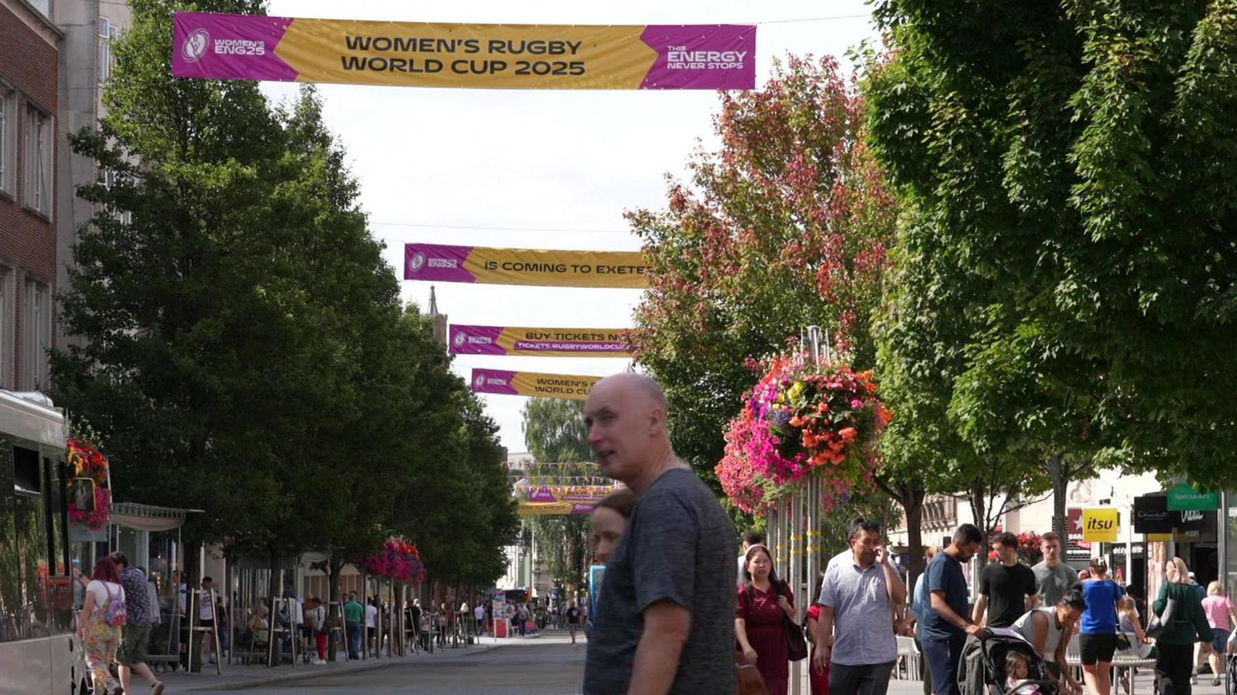 People walk around a busy Exeter High Street. There are pink and yellow banners hoisted up over the street advertising the 2025 Women's Rugby World Cup. Trees and hanging baskets with colourful flowers line both sides of the road.