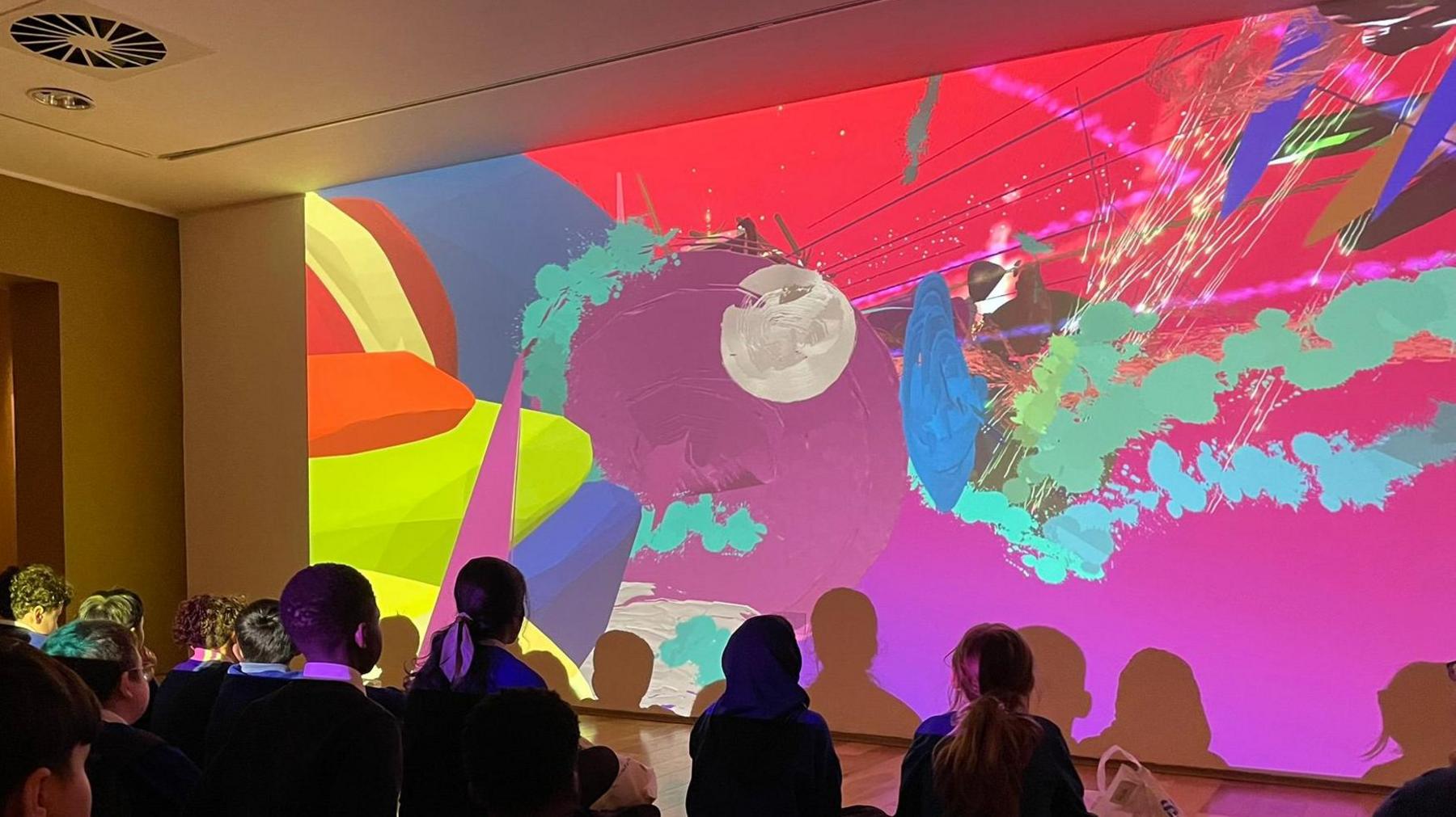 A group of schoolchildren looking at brightly coloured artwork that has been projected onto a wall. They are sitting on the floor, with their backs to the camera. You can just see the backs of their heads and their upper bodies. The artwork is in red, yellows, blues and purples and shows lots of different shapes. 