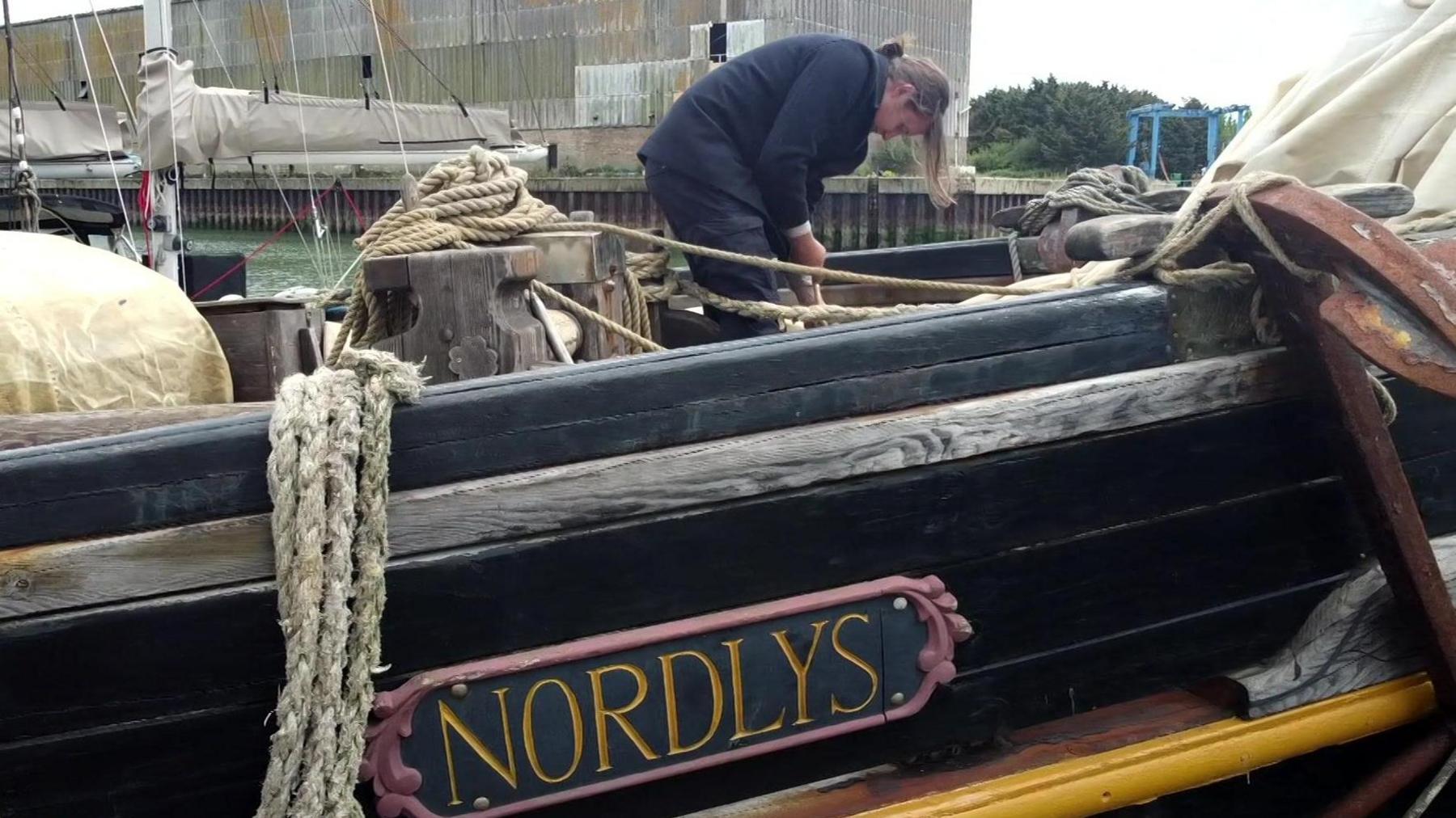 The side of the Nordlys ship is painted black with a sign that says the boat's name in gold. William Lund is standing on the boat's deck, bending down looking at rope.