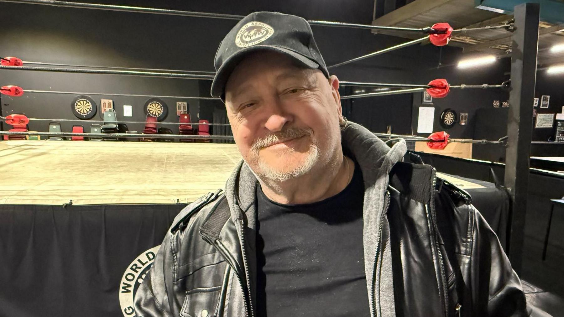 Wrestler Ricky Knight stood beside a ring smiling. He has a grey goatie beard and is wearing a black jacket and baseball cap