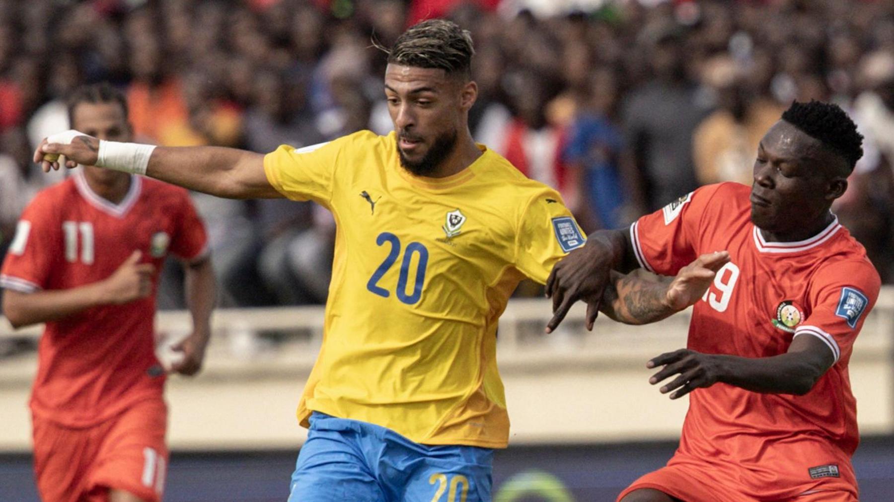 Denis Bouanga wears a yellow Gabon shirt with a number 20 on the chest and blue shorts as he holds out his arms to balance and shield the ball during a football match from Kenya defender Rooney Onyango, who is wearing a red shirt. A second Kenya player can be seen in the background running towards the pair while behind him you can see a blurred packed stand full of supporters
