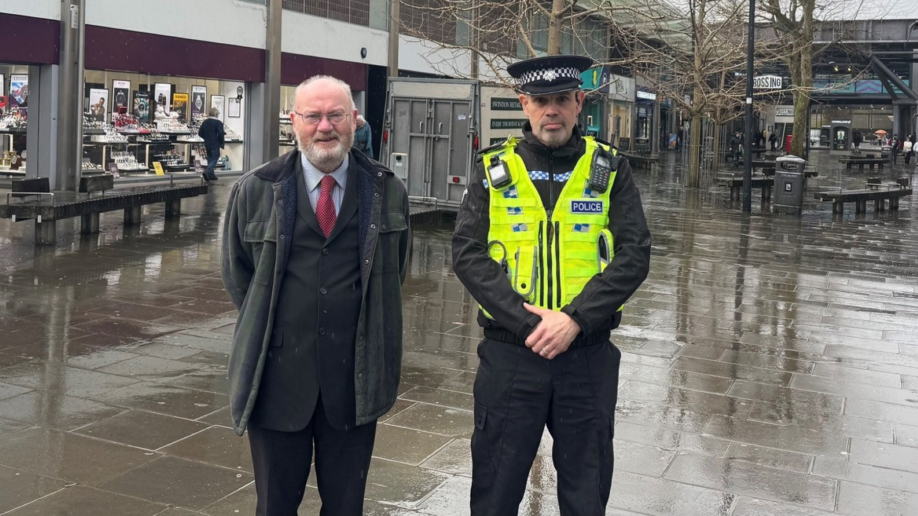 Two men face the camera, one in a suit and tie, the other in a police uniform in a town centre with shops, benches, trees and a public bin in the background, which appears wet