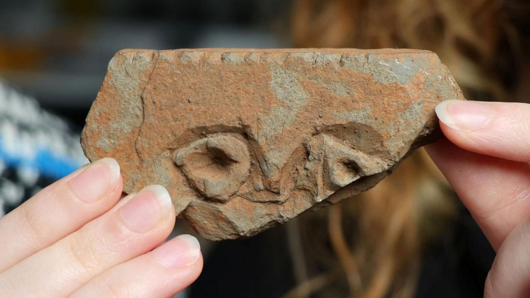 The wedge-shaped stone is a rusty red colour. There are carved notches along the flat top of the stone, and shapes of two eyes. The stone is being held in the hands University of Aberdeen student Jodie Allan.