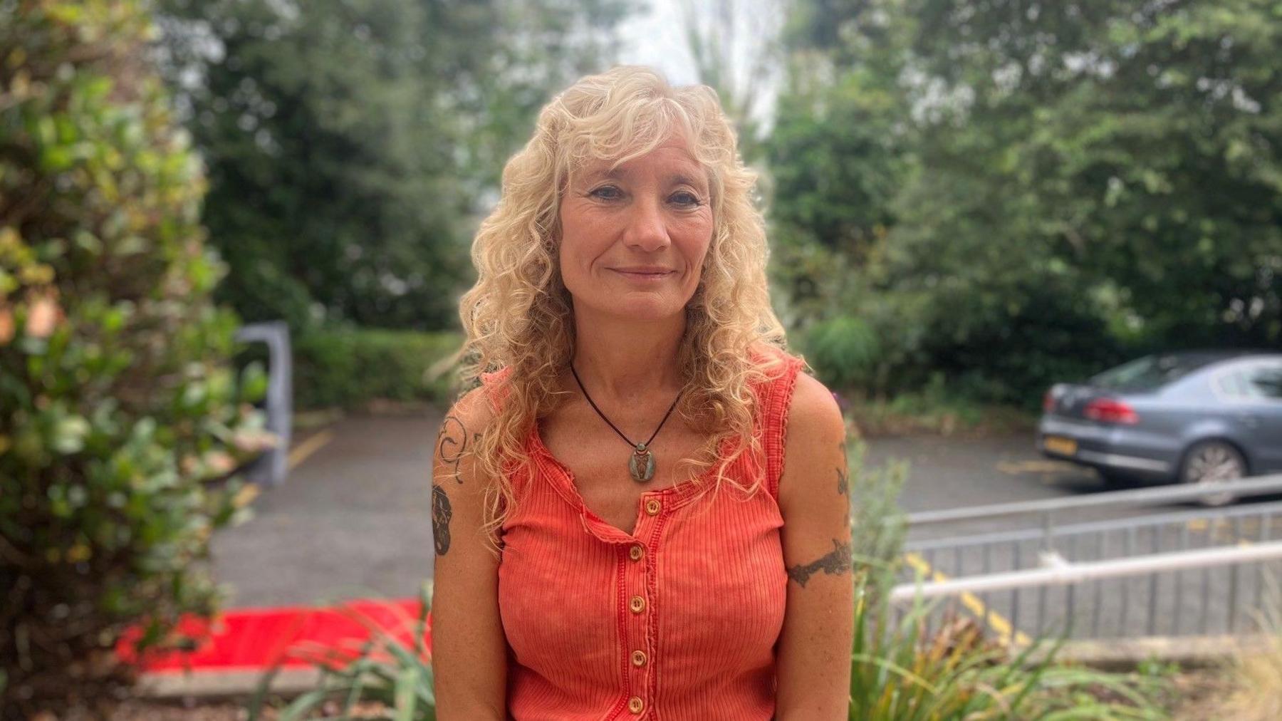 A woman sits outside the Hospital smiling . She has blonde curly hair and wears a red vest top 