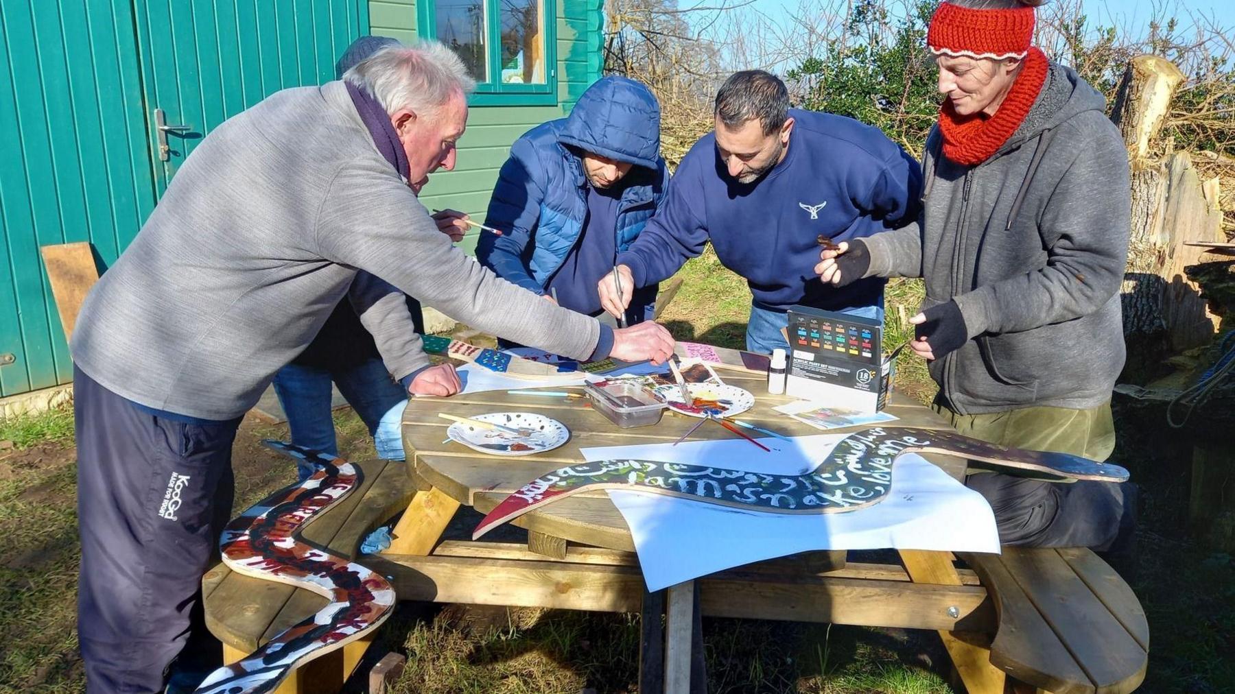 Five volunteers wearing tracksuit bottoms, jumpers, hats and coats. They are leaning over a round wooden picnic table and painting large wooden snakes. On the table there are various paints, a mixing palette and a Tupperware used to wash paintbrushes. They are outside on a cold but sunny day, and in the background there is a large green wooden shed. 