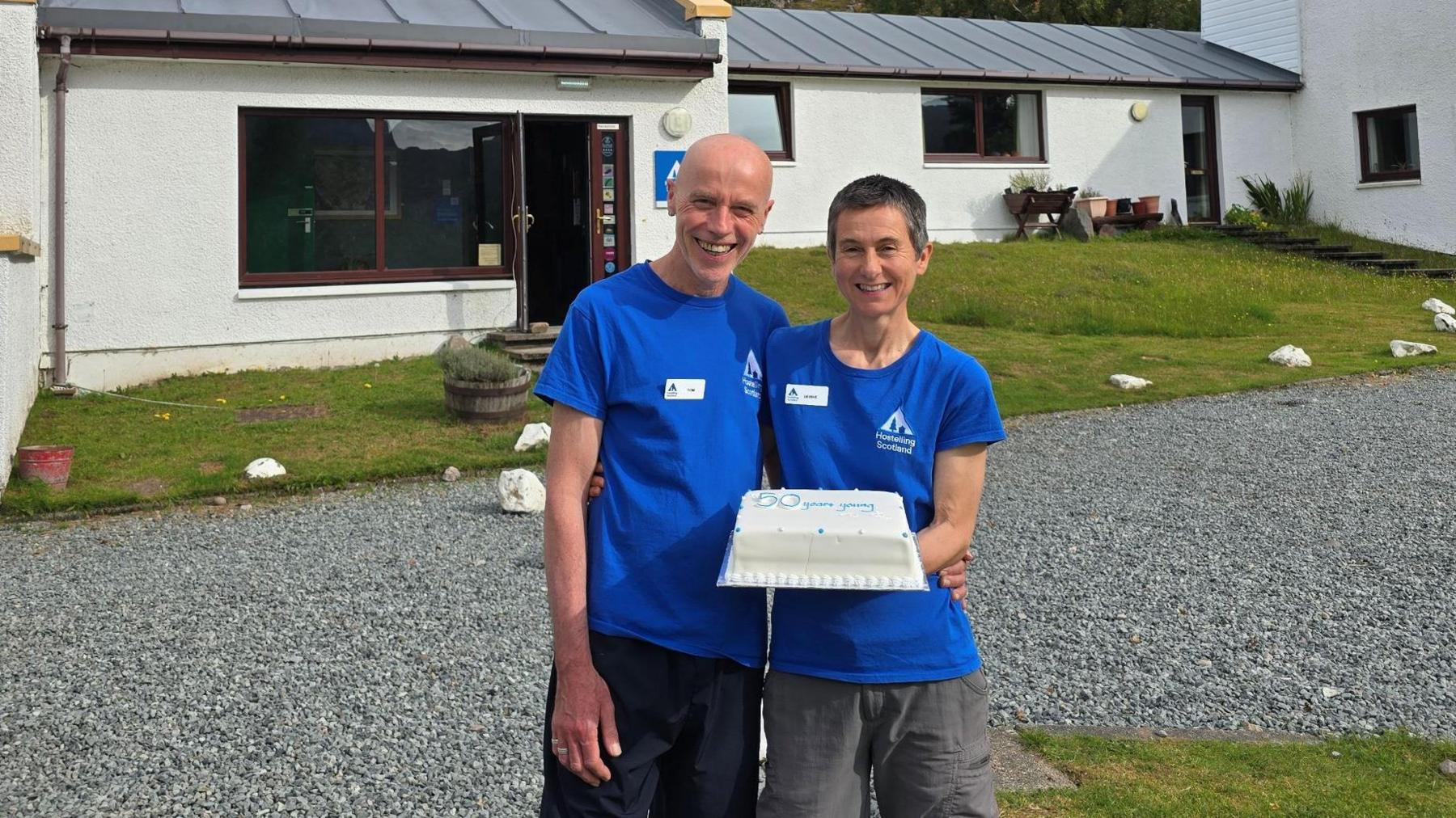A man with a bald head and a woman with short dark hair stand next to each other with the cake outside the youth hostel. The cake has white and blue icing. The youth hostel is made up of a number of white-walled, grey roofed buildings. A mountain rises behind the hostel.