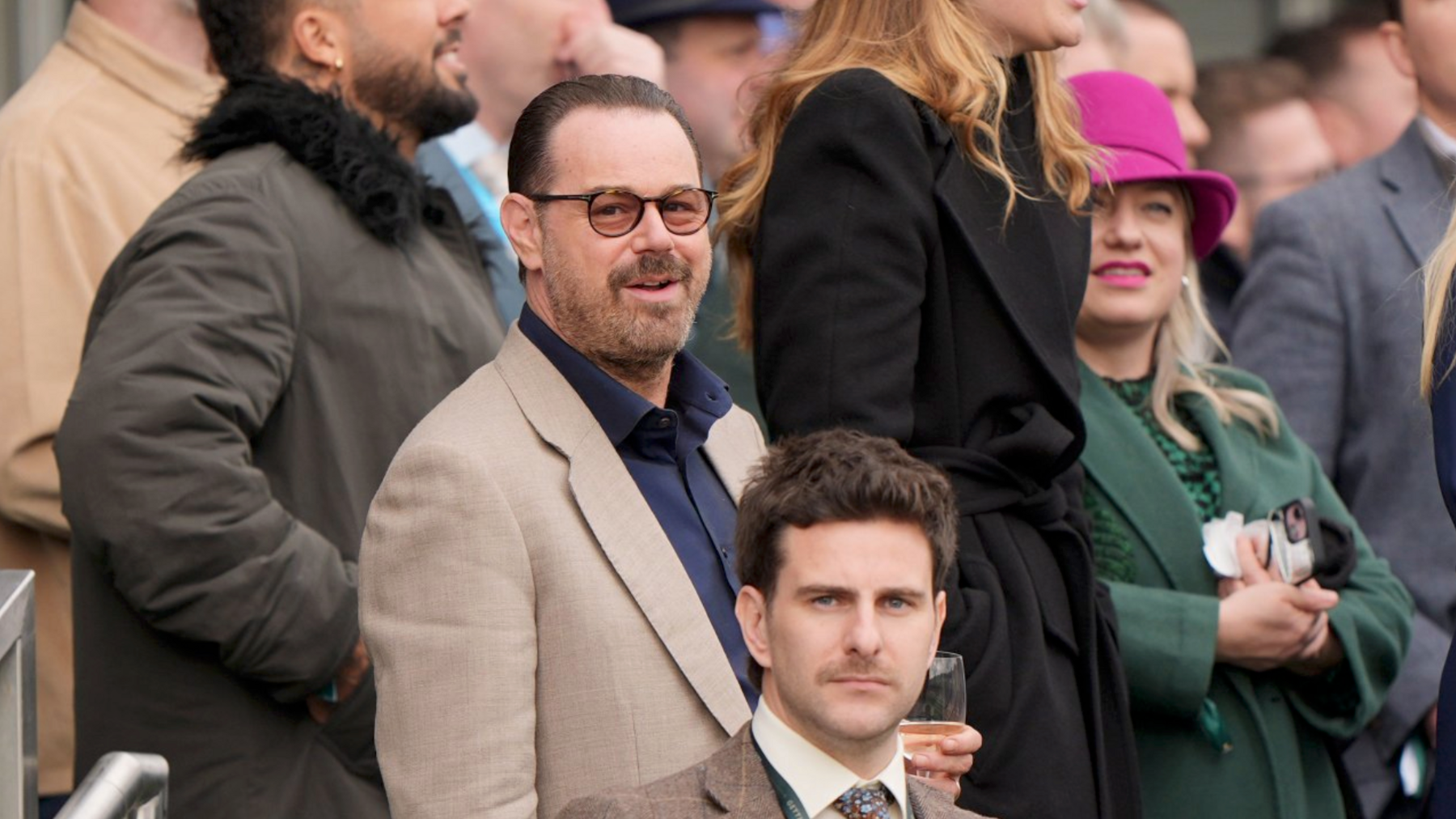 Actor Danny Dyer is smiling while wearing a brown blazer over a dark blue shirt. He has black-rimmed glasses with orange lenses and is holding a glass. He is standing in a crowd of people.