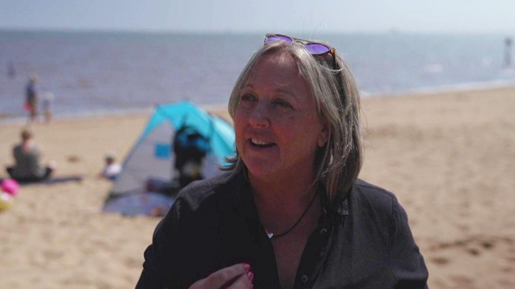 A woman with shoulder-length hair and sunglasses on her head. She is wearing a necklace and a dark-coloured blouse and is stood on a beach. A blue tent and people sitting on the sand can be seen behind her. The sea is in the distance.