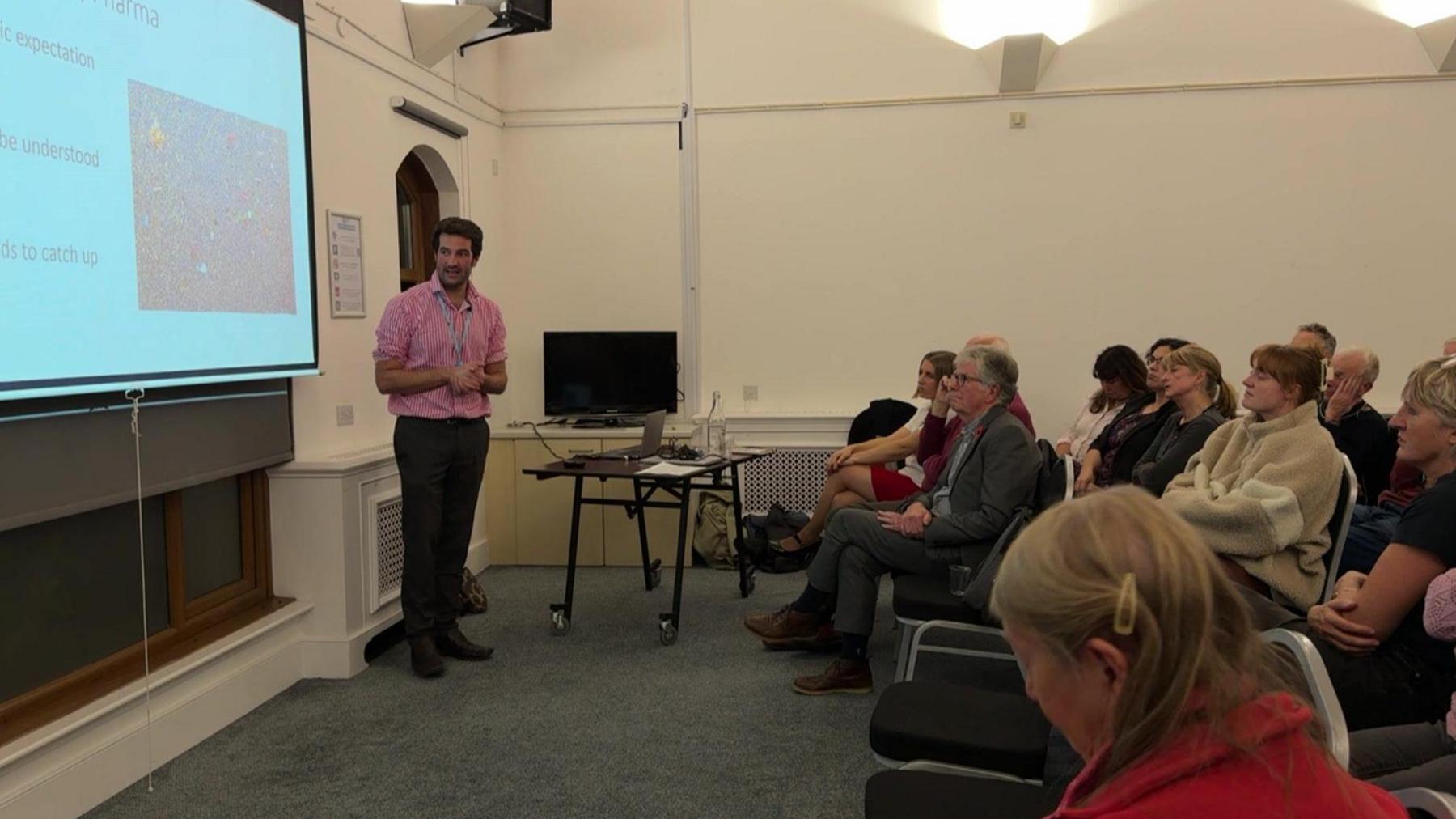 A wide view of the public meeting with a man in a pink shirt standing beside a projector screen with an audience watching