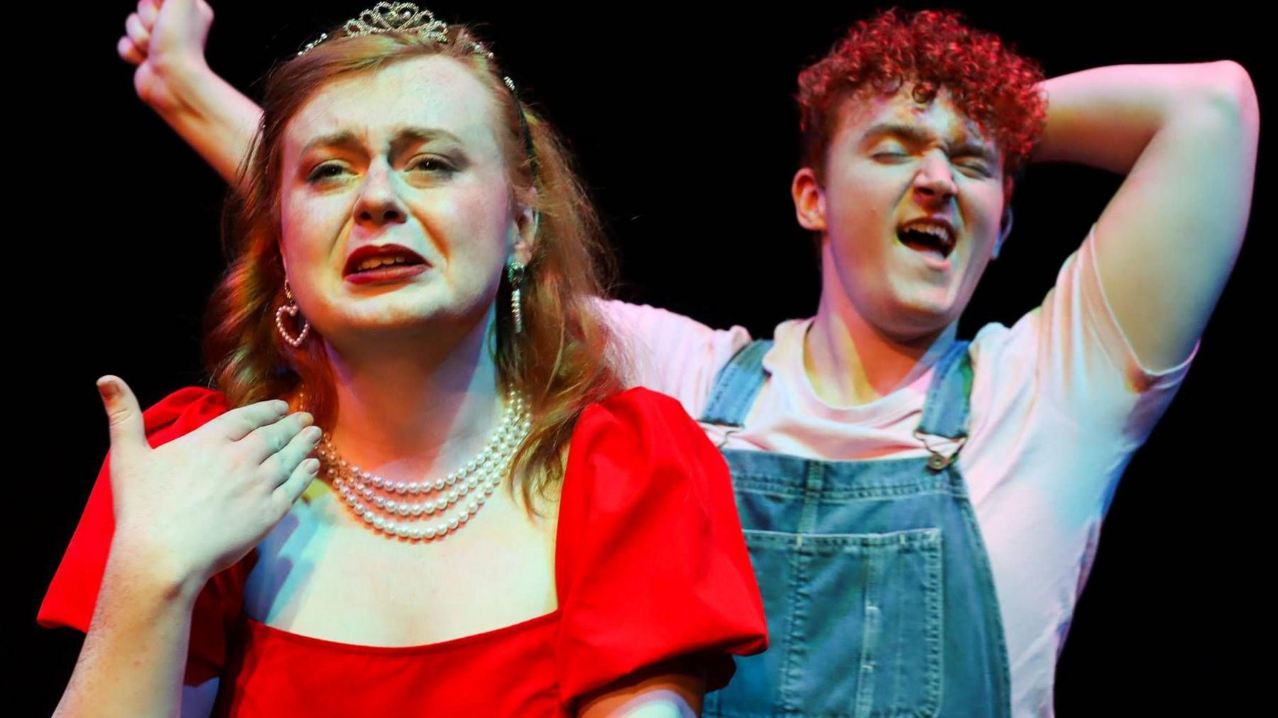 Rose and Conor, with a pitch-black background behind them. Rose is wearing a red dress, while Conor is wearing a white t-shirt and blue overalls. 
