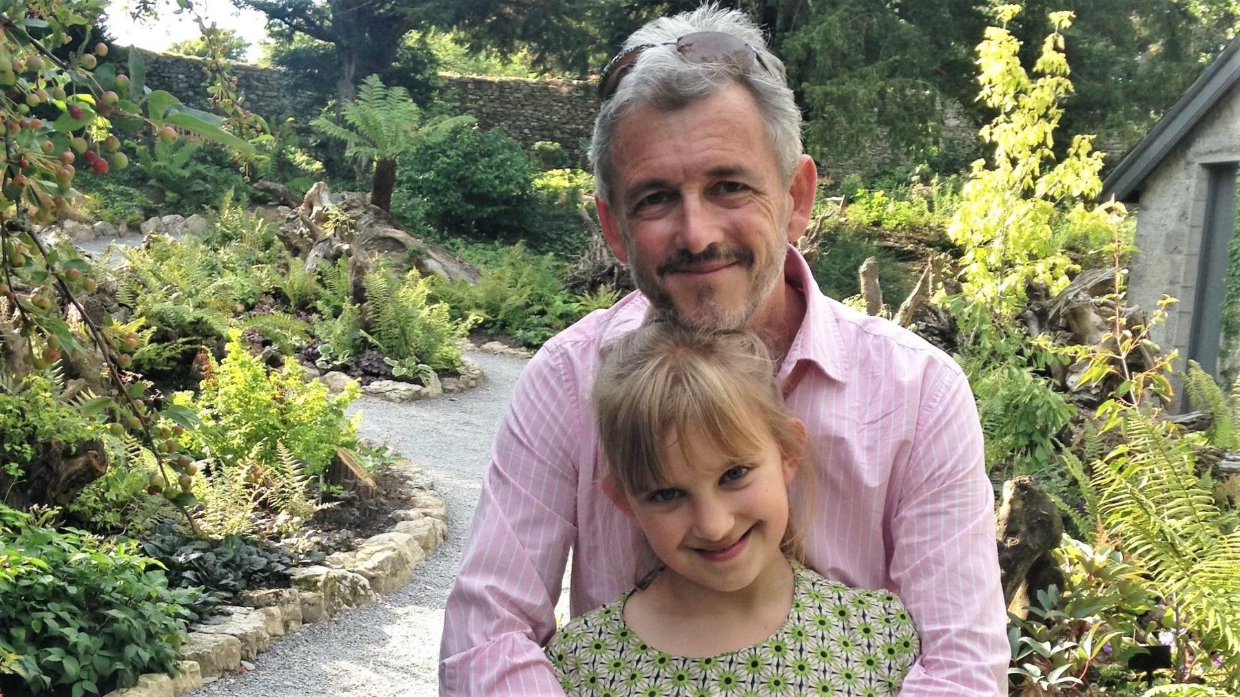 Mike Calnan with his young daughter Flossy. He is wearing a pink pinstripe shirt with sunglasses on his head, and is smiling at the camera with his arms wrapped around Flossy's shoulders. She is wearing a green patterned top and has her blonde hair tied back into a pony tail with a fringe. They are standing in a garden on a sunny day, surrounded by lots of greenery and winding gravel paths.