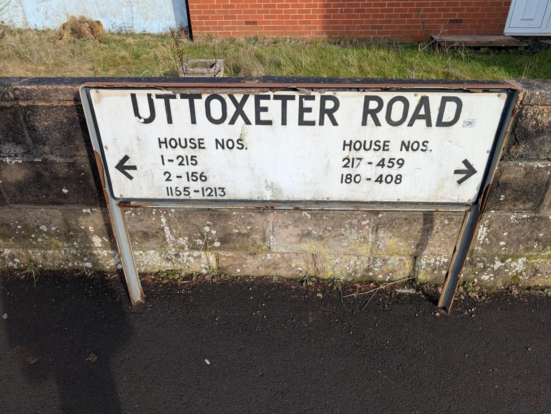 A white, horizontal street sign with black letters saying Uttoxeter Road. It is suspended by two metal posts and stands against a shallow brick wall, which has some grass and a residential property behind it. A black arrow icon at each end of the sign points in opposite directions. Alongside each arrow are house numbers, properties to which the arrows point 