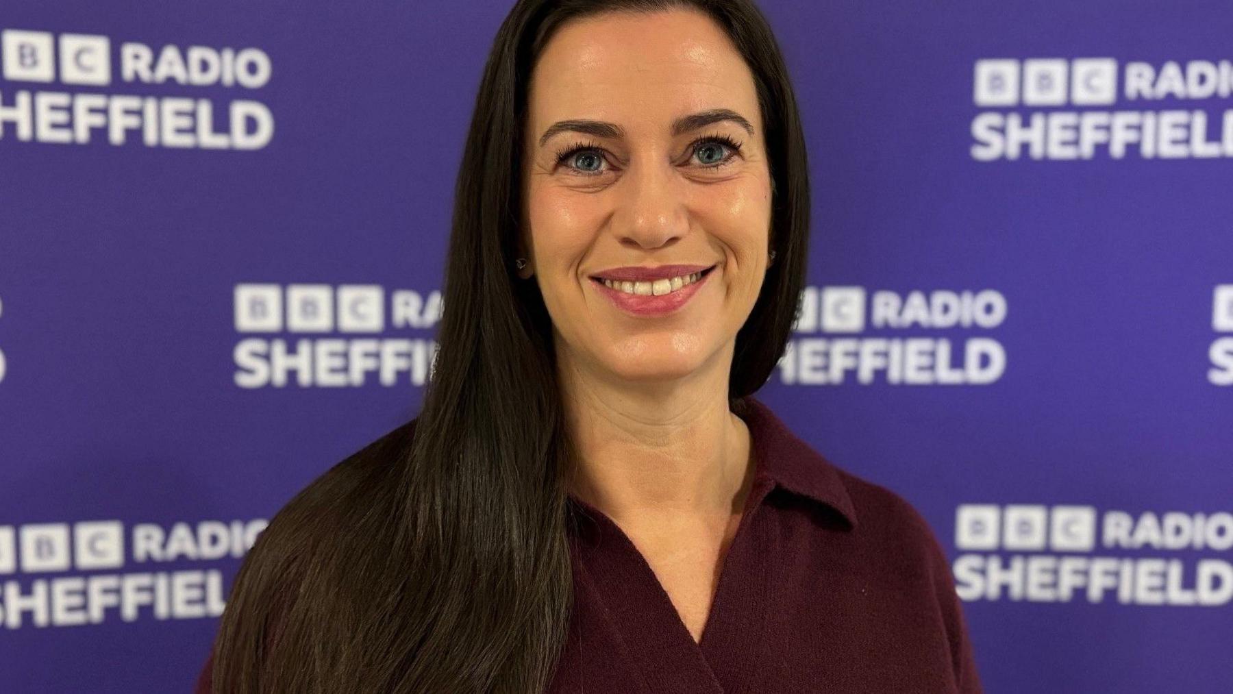 Tracy Richardson at BBC Sheffield, November 2025, wearing a plum coloured jumper with long black hair and the purple BBC Sheffield background