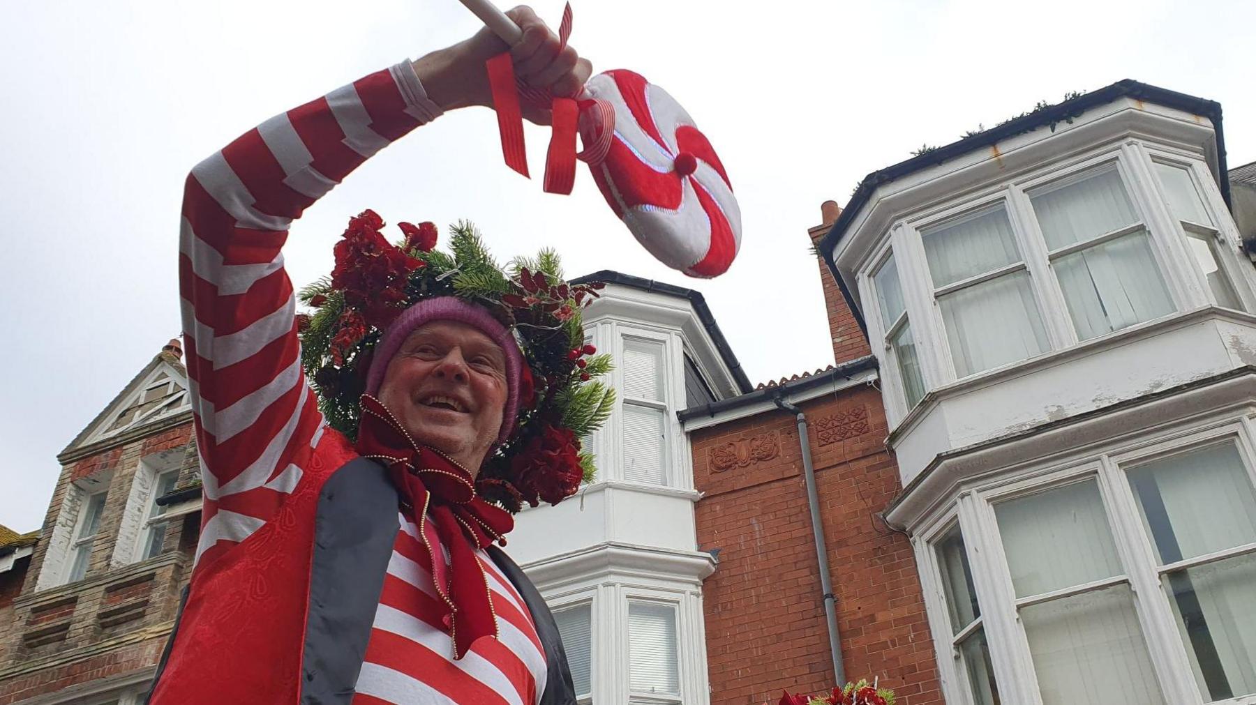 A male stilt walker dressed as an elf. The camera is looking up at him as he smiles and waves to the crowd.