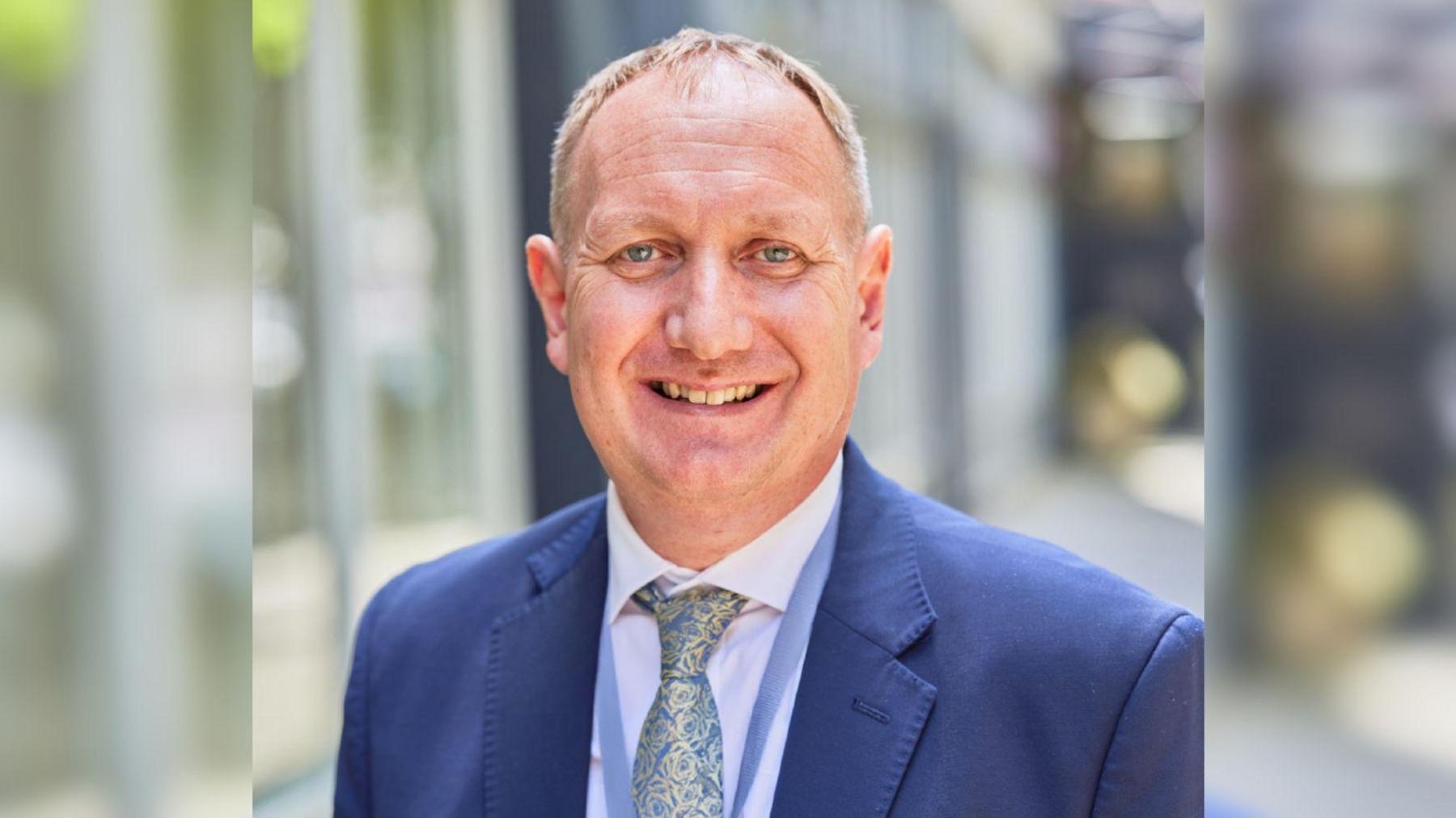 Andy Berry wearing a blue suit, white collared shirt and blue and gold floral patterned tie. It is a professional portrait style photo, and he is smiling at the camera with a soft blurry background. 