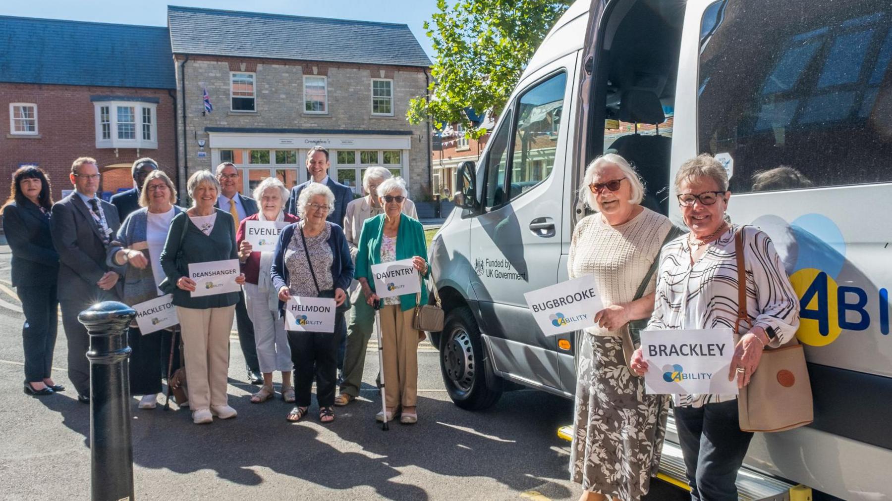 Group of people holding signs with names of different south Northamptonshire towns and villages pose next to the Ability community bus