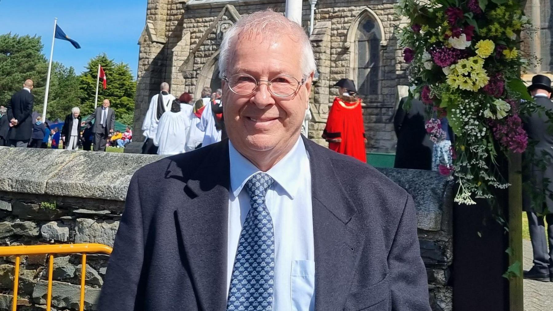 A smiling Trevor Nall wears a blue suit and light blue shirt with a blue tie. A crowd of clergy and officials gather in the background in front of the Royal Chapel in St John's.