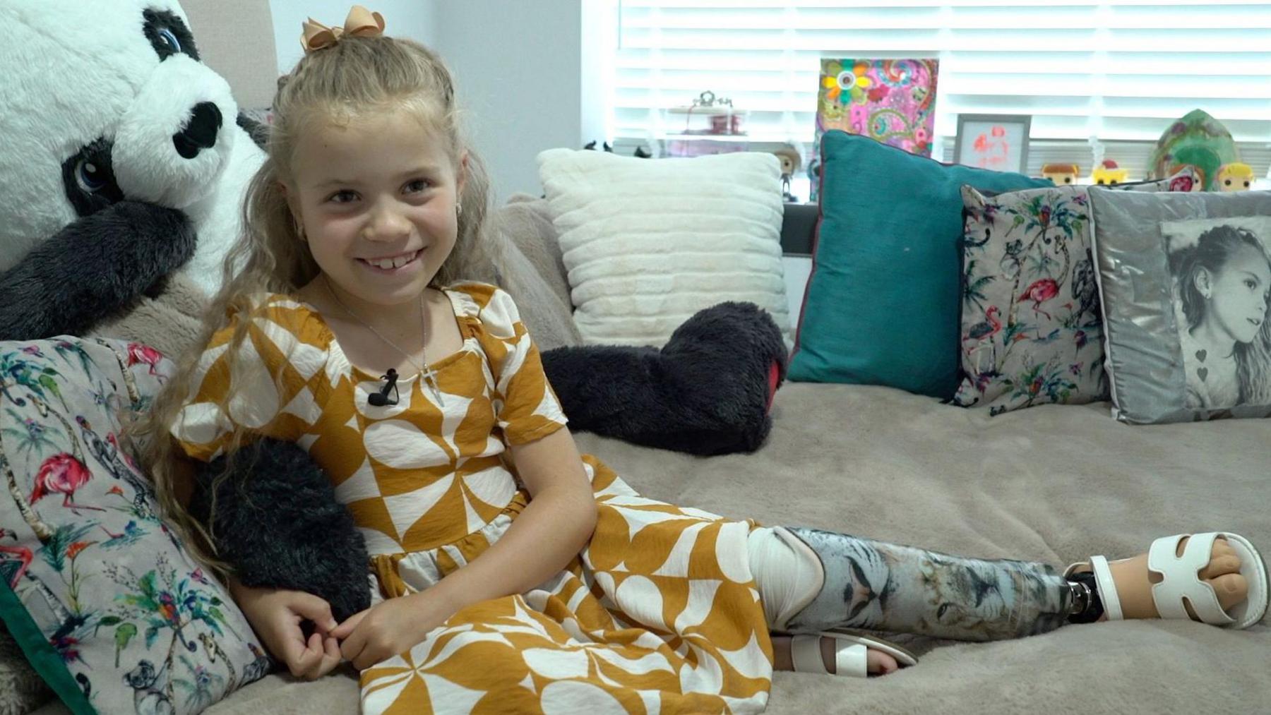 A young girl sits on a light grey couch. She is surrounded by cushions and wears a mustard and white patterned dress and has blonde hair.