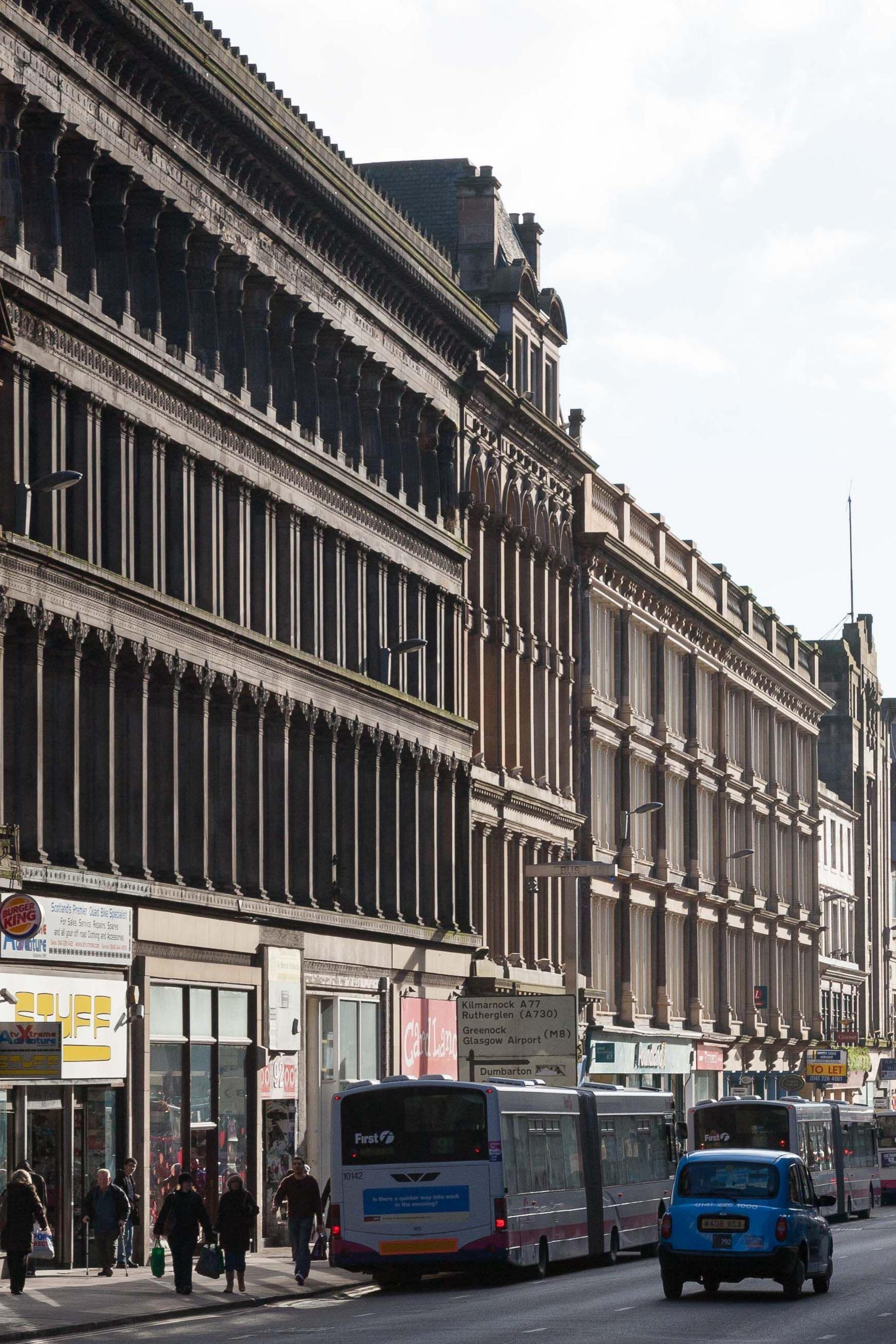 Side view of the Egyptian Halls, a wide sandstone building with decorative pillars. Busses, a taxi and pedestrians are on the street below.