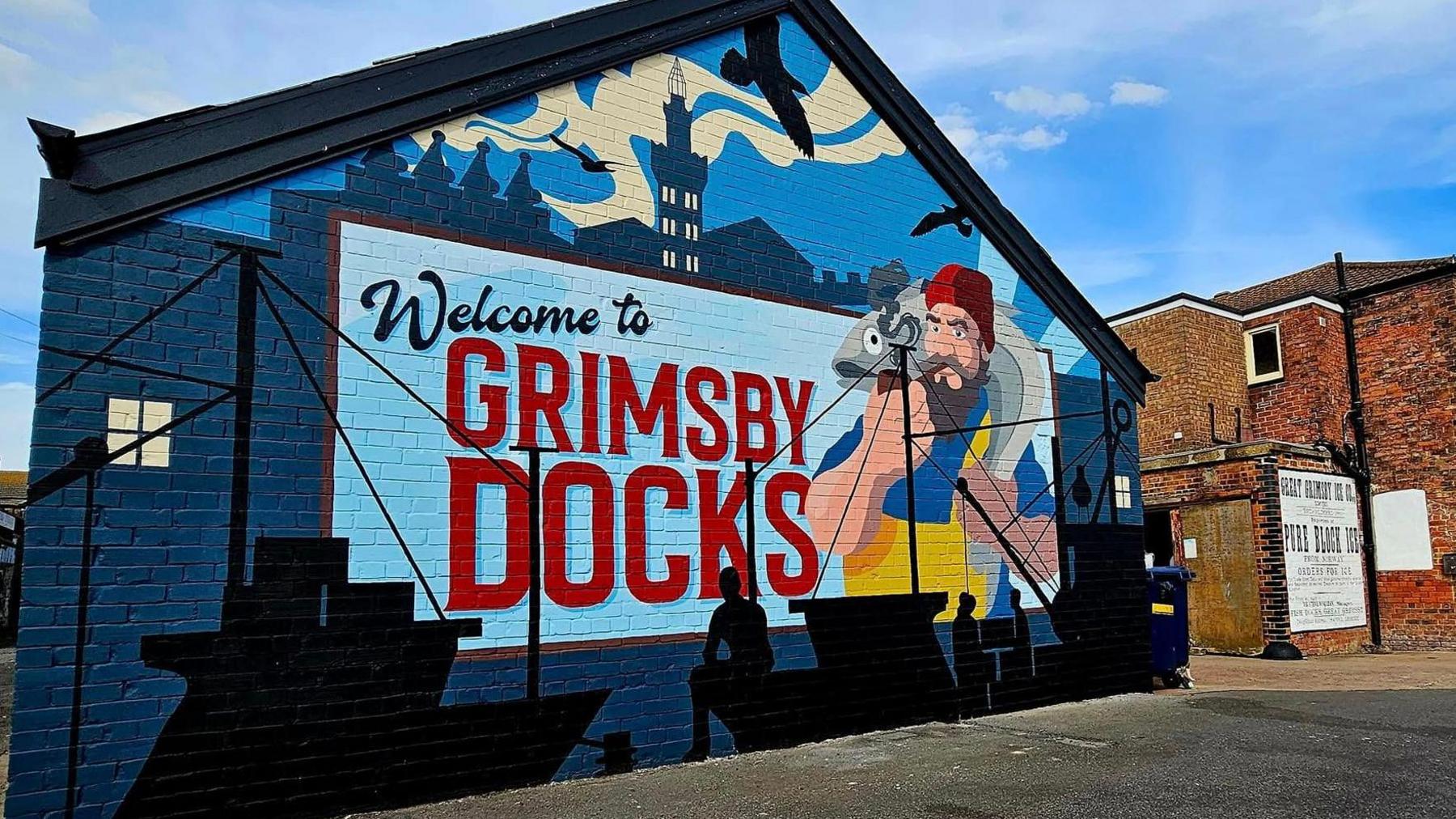 A large blue painting displayed on a brick wall. The mural details the words: "Welcome to Grimsby Docks" in bold black and red writing. Alongside the words- there is a bearded fisherman holding a large fish to the right. There are also silhouettes of boats and trawlers at the bottom of the wall. The mural is next to a red bricked building with a white and black poster on- the words are illegible. 