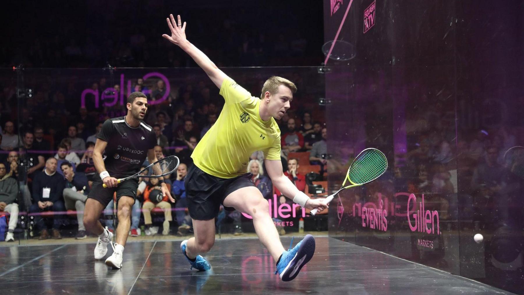 In a squash court with an audience, a man is about to swing a squash racket while wearing a yellow and black sports kit and blue trainers. He is trying to hit a white squash ball before it bounces on the floor. Behind them there is a another player looking on.