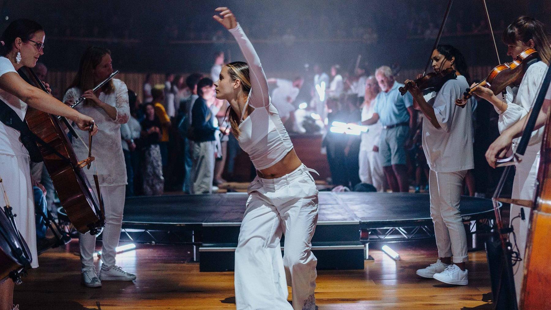 A young woman all in white dances between rows of musicians, also all in white, as part of the Paraorchestra event during the BBC Proms at Bristol Beacon