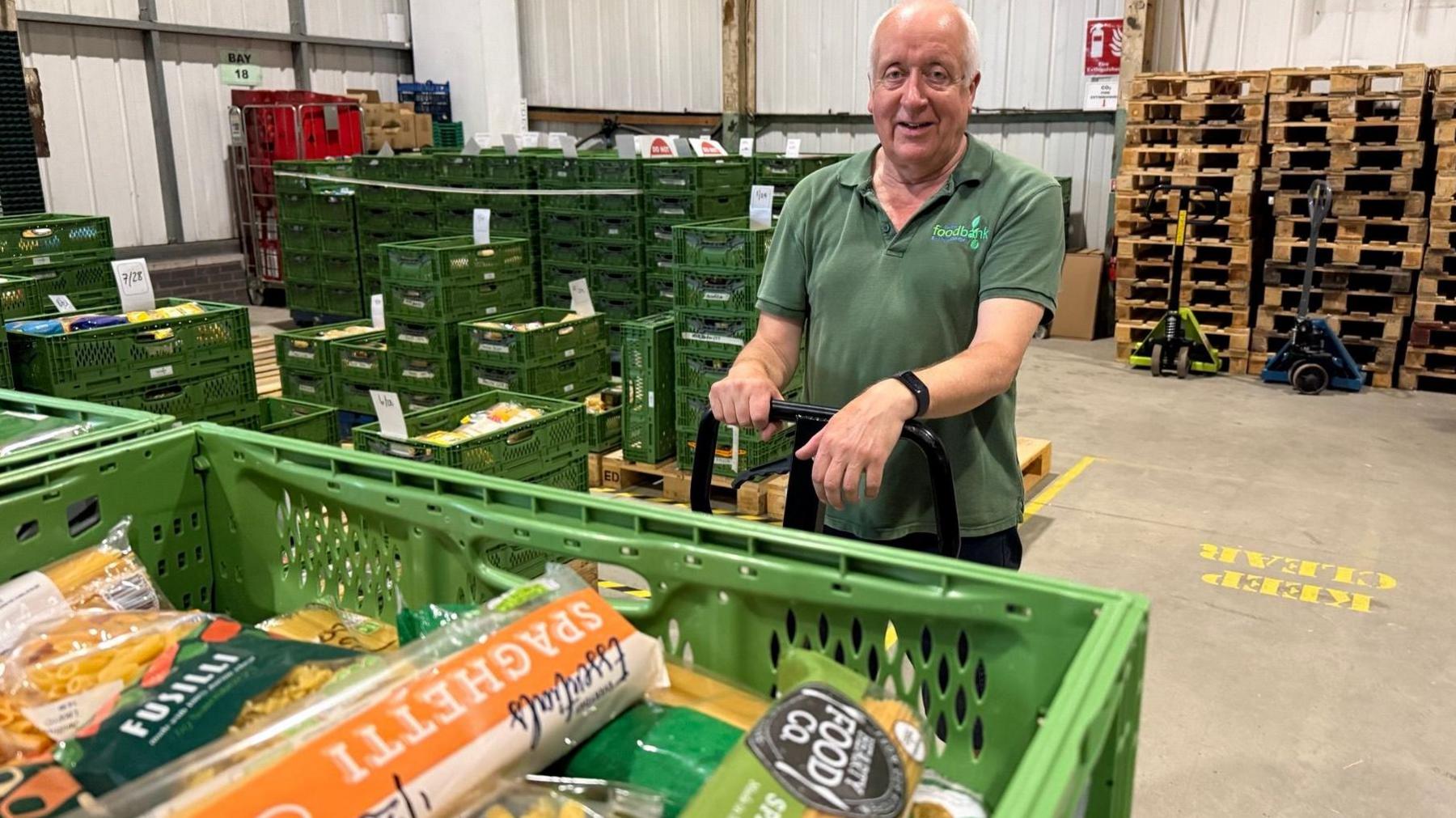 Volunteer, Paul Twyman, wears a green food bank polo shirt and stands in front of a pallet of pasta which has just been delivered. 