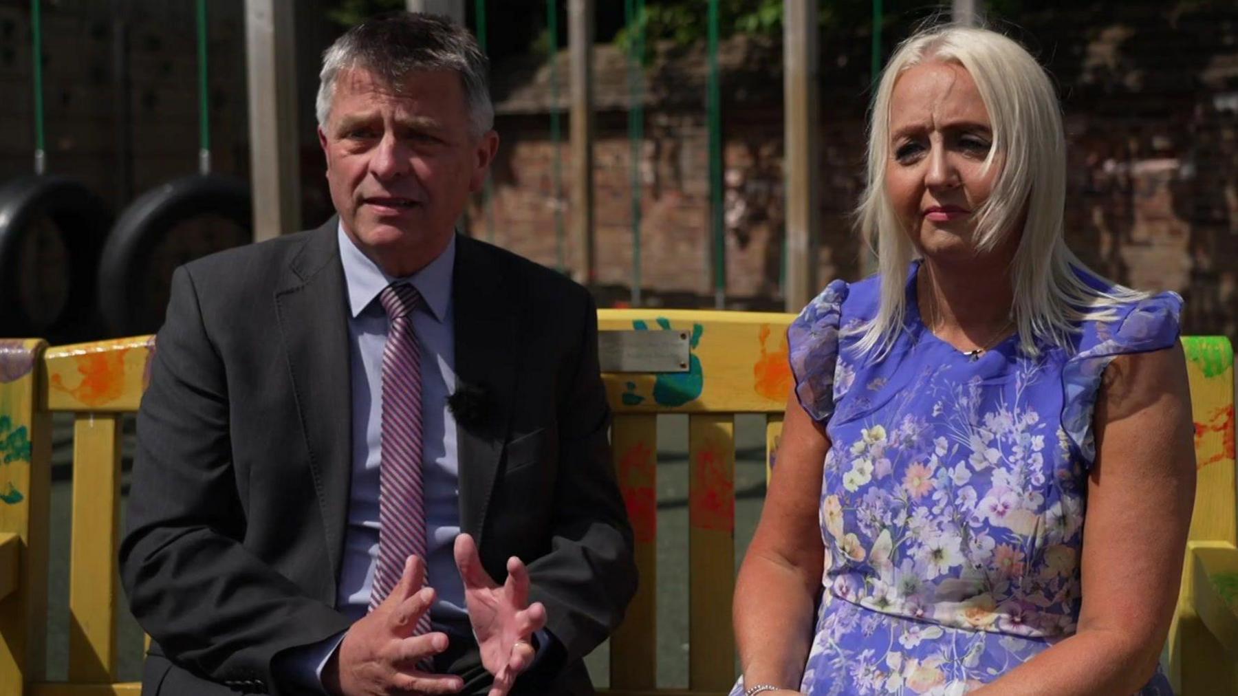 A wide shot of Adrian Antell and Jennie Sephton. Adrina has short grey hair and is wearing a dark grey suit with a striped tie and light blue shirt. Jennie has blonde hair and is wearing a blue flowery dress.