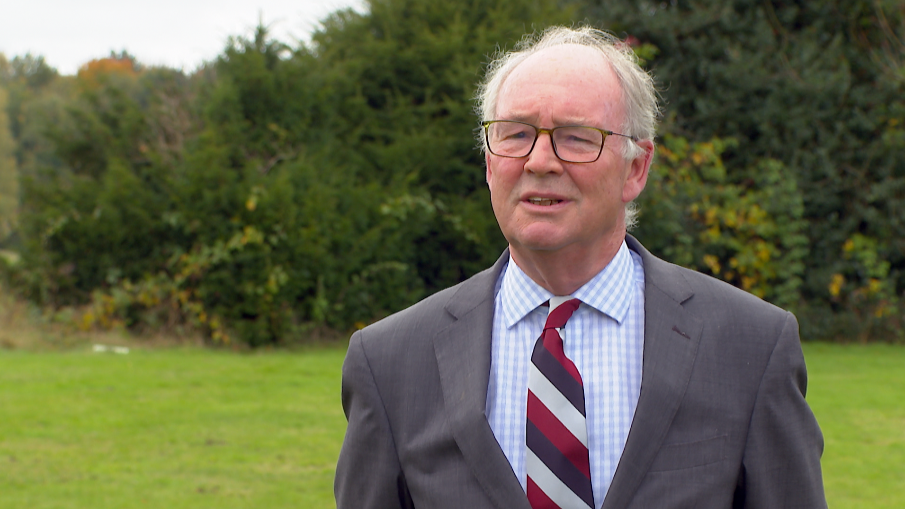 A man wearing glasses and a grey suit with a blue and white cross hatched shirt and a red, navy and white striped tie. He is stood outdoors with an area of grass and trees in the background.