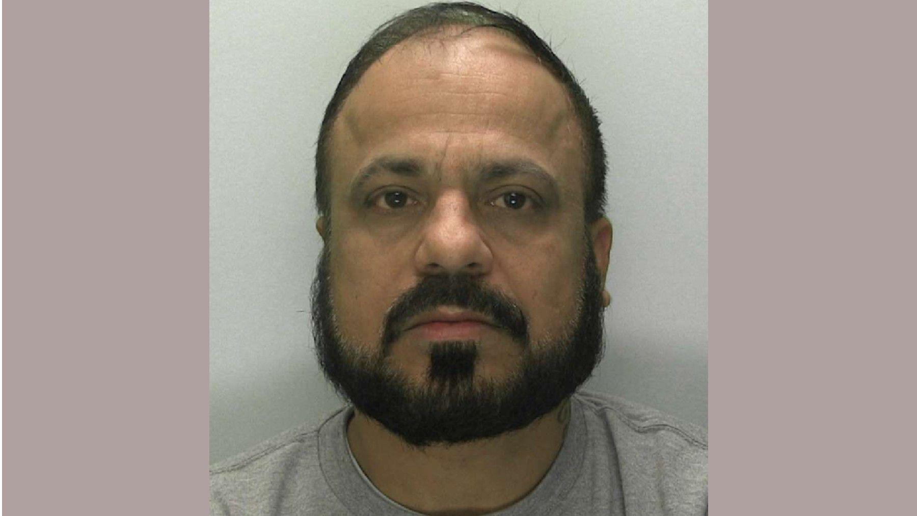 A custody mugshot of a middle-aged man with short, thinning black hair, black facial hair in the form of a short goatee beard and moustache, with a blank expression. He is wearing a grey top.