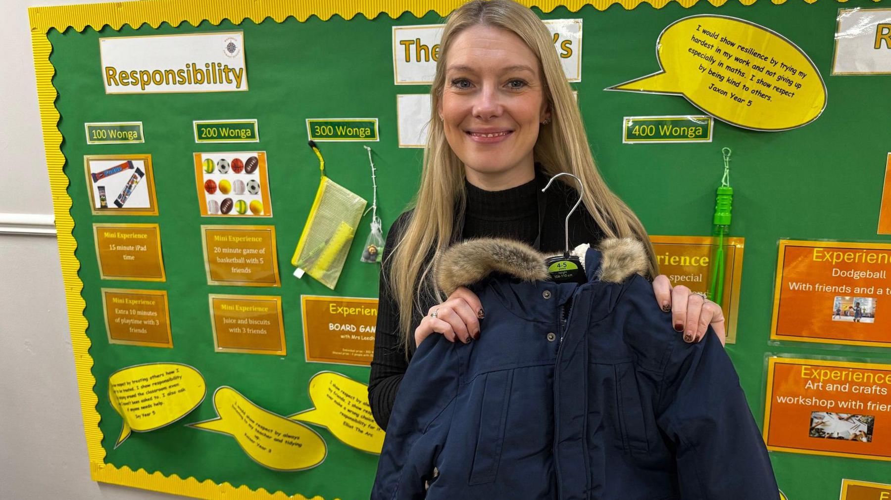 A woman with long blonde hair holds up a navy blue Parka-style coat. She is standing in a school corridor in front of a colourful wall display, which has headings such as "Responsibility"