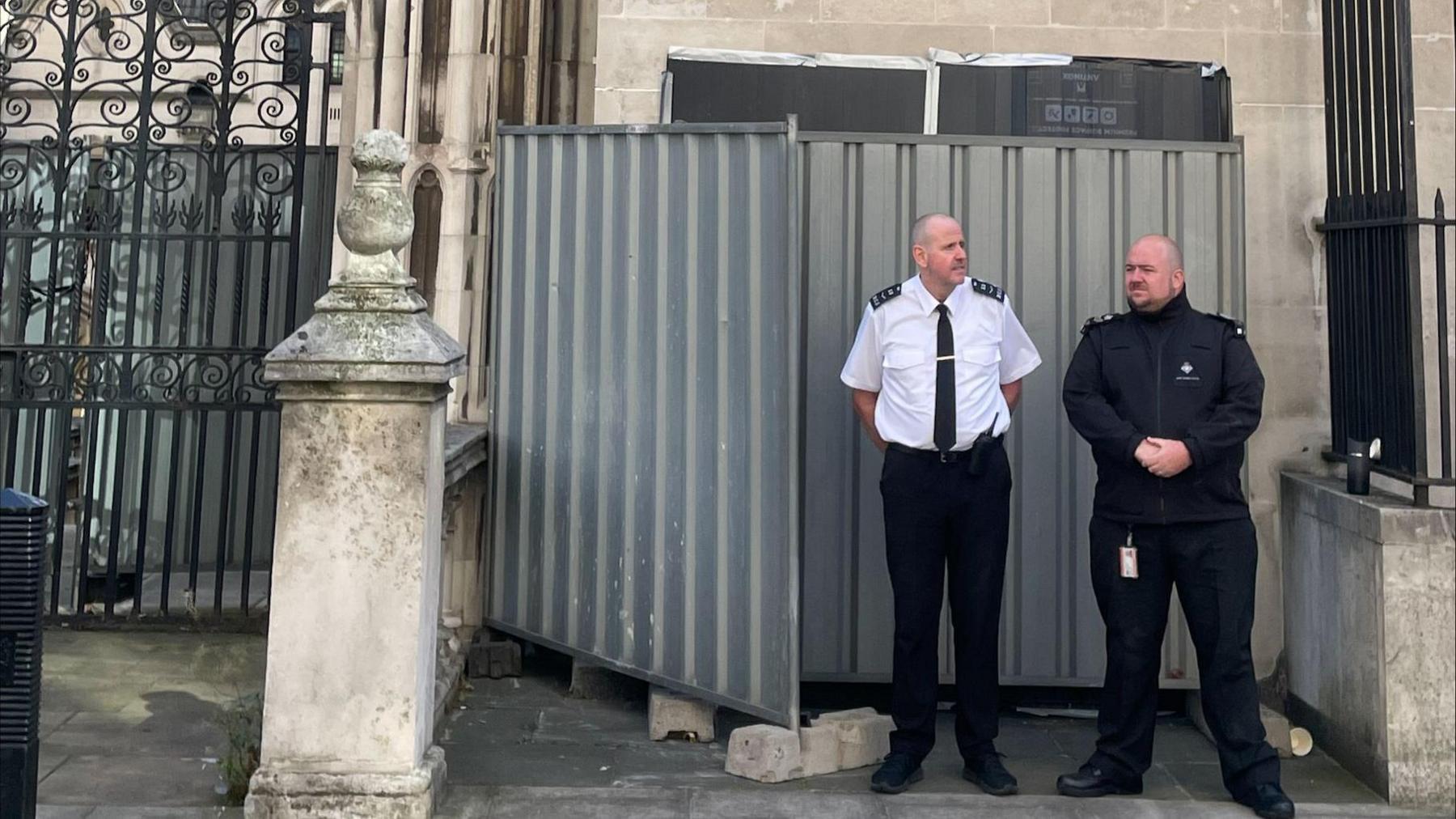 Two security guards at the Royal Courts of Justice, standing in front of grey metallic barriers blocking the Banksy artwork from view. High above on the wall is a CCTV camera.