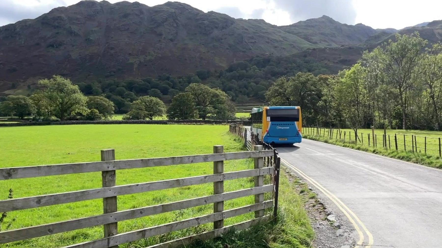 A small road with a Stagecoach bus being driven away, with a backshot of the Lake District fells