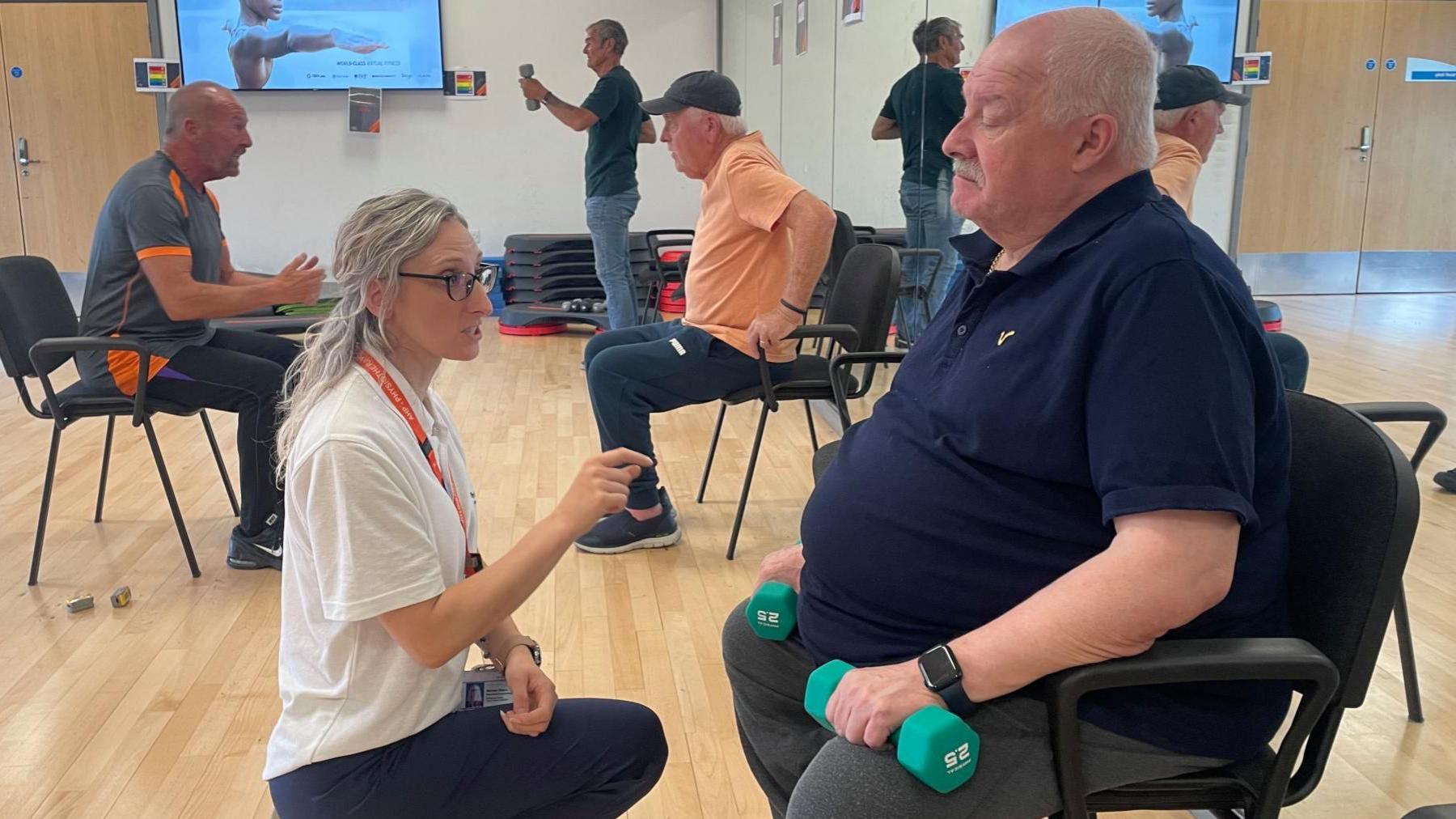 A woman with long blonde hair crouches down to encourage an older man sitting in chair with small green weights. In the foreground other men of the gym other men do stretching exercises