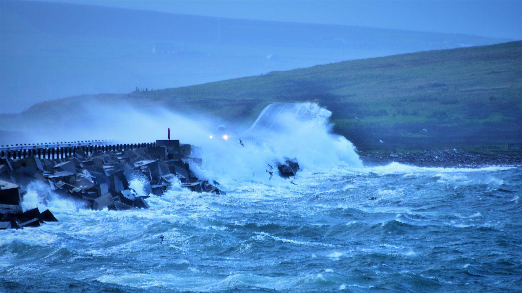 Headlights of a car approaching the causeway in Orkney in stormy weather. Strong waves are crashing over the road. 