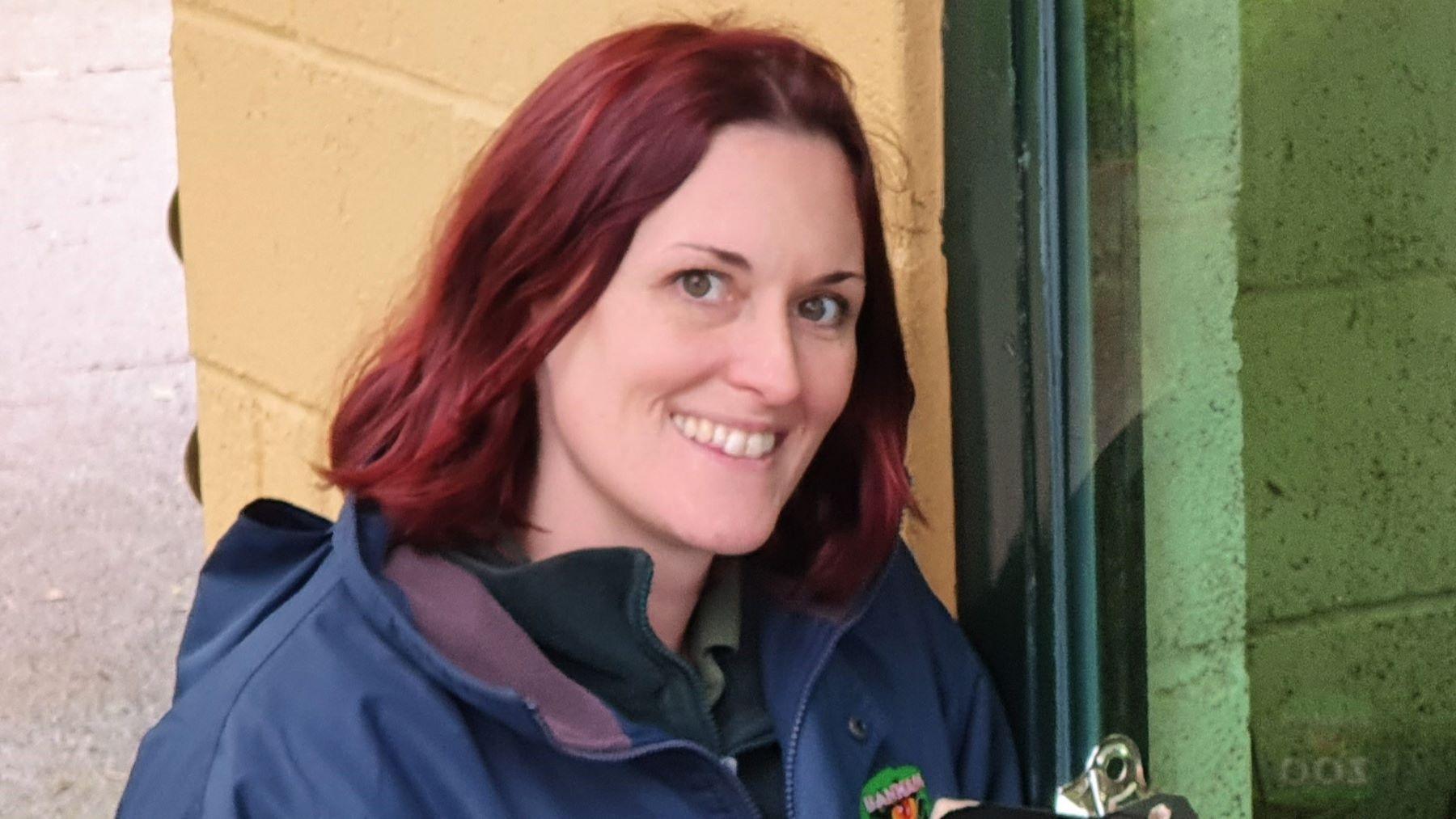 Deborah Harris crouched by a yellow wall. She is wearing a blue jacket with a Banham Zoo logo. She has shoulder-length red hair and is smiling.