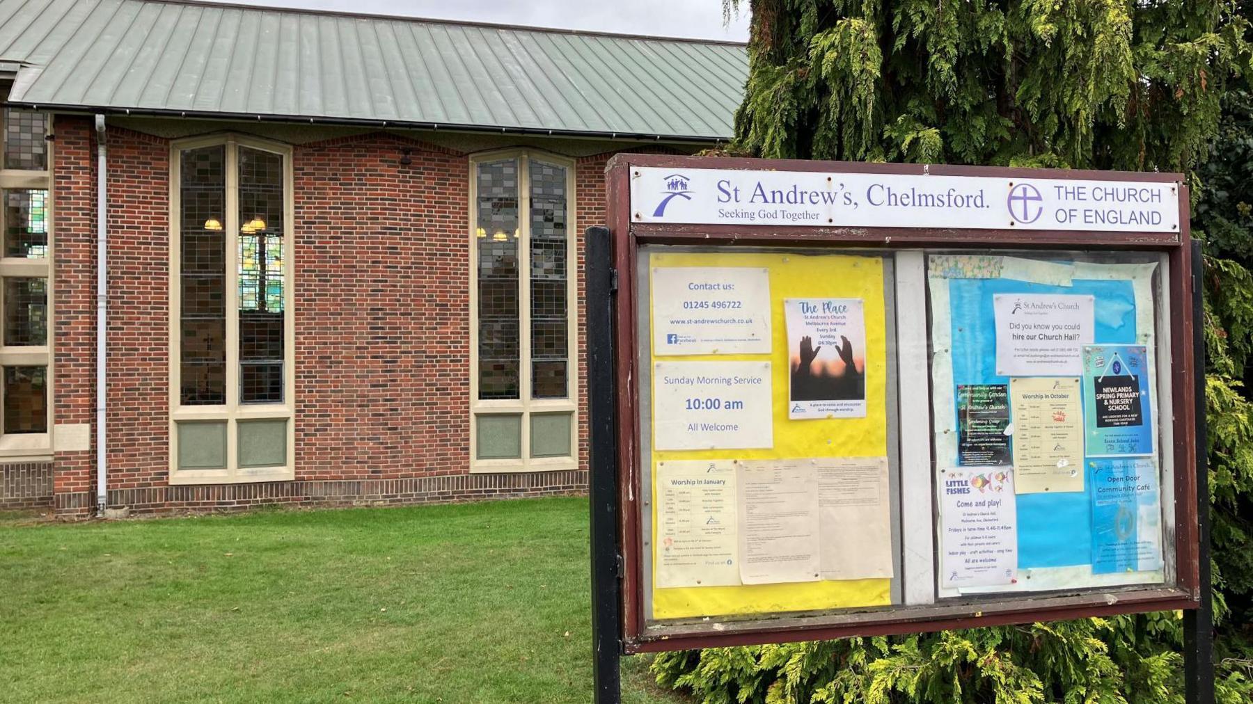 The church is a large brick building with a grey roof and tall stained glass windows. In the foreground is a wooden notice board advertising its services.