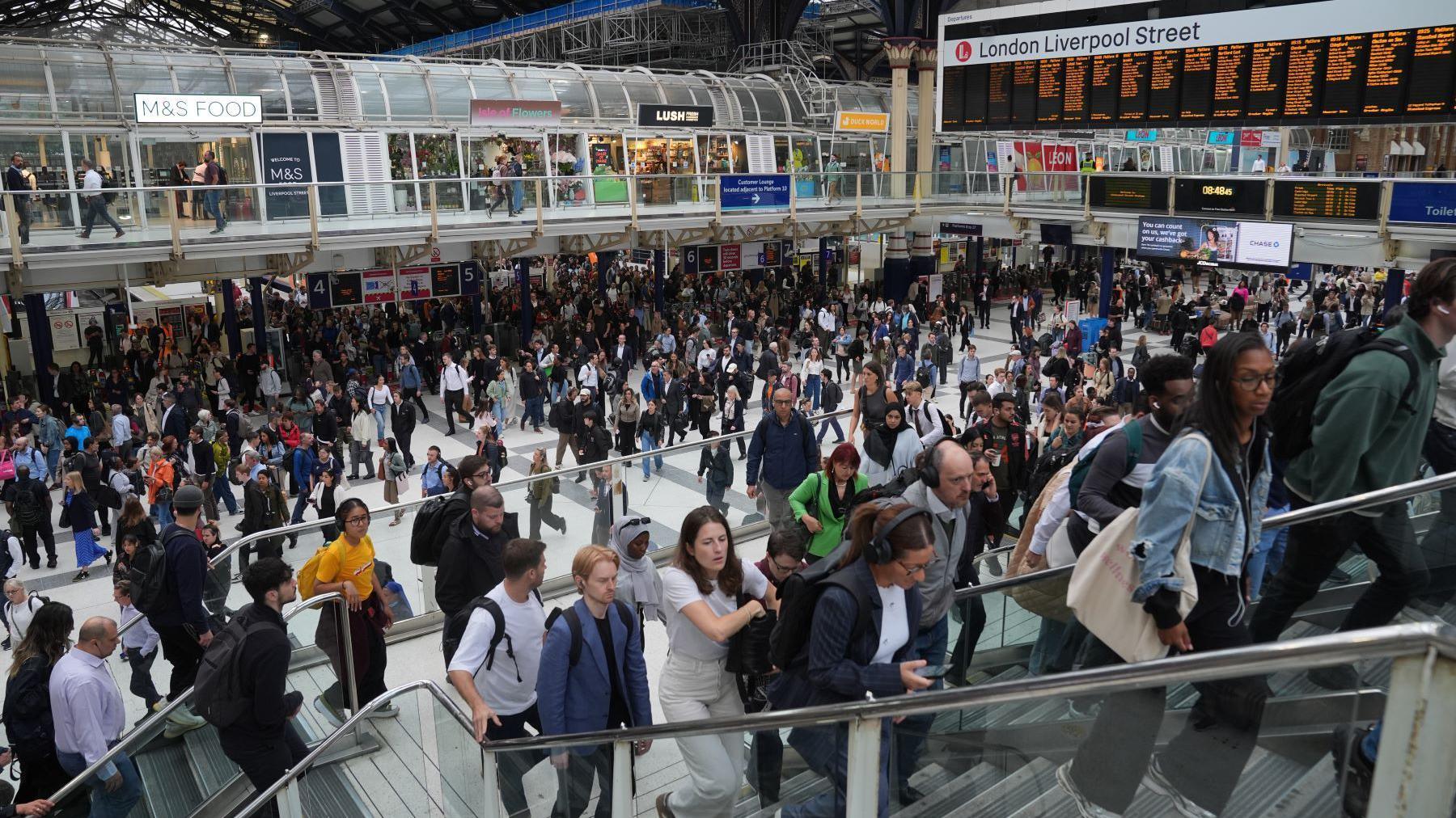 Hundreds of people, if not thousands, are crowding and walking through the main hall of London Liverpool Street. In the background are the digital departure screens. In the foreground people walk up stairs.