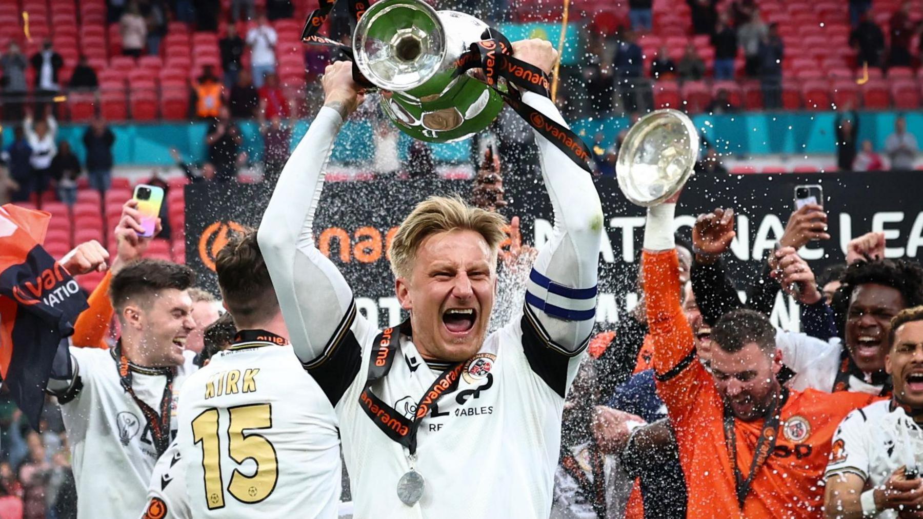 Byron Webster holds the National League promotion final winners' trophy aloft cheering, while teammates around him celebrate 