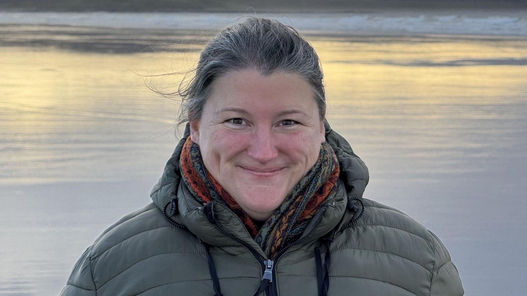 Lucy Cowley yn sefyll o flaen llyn. Mae hi'n gwisgo cot werdd ac edrych tuag at y camera.