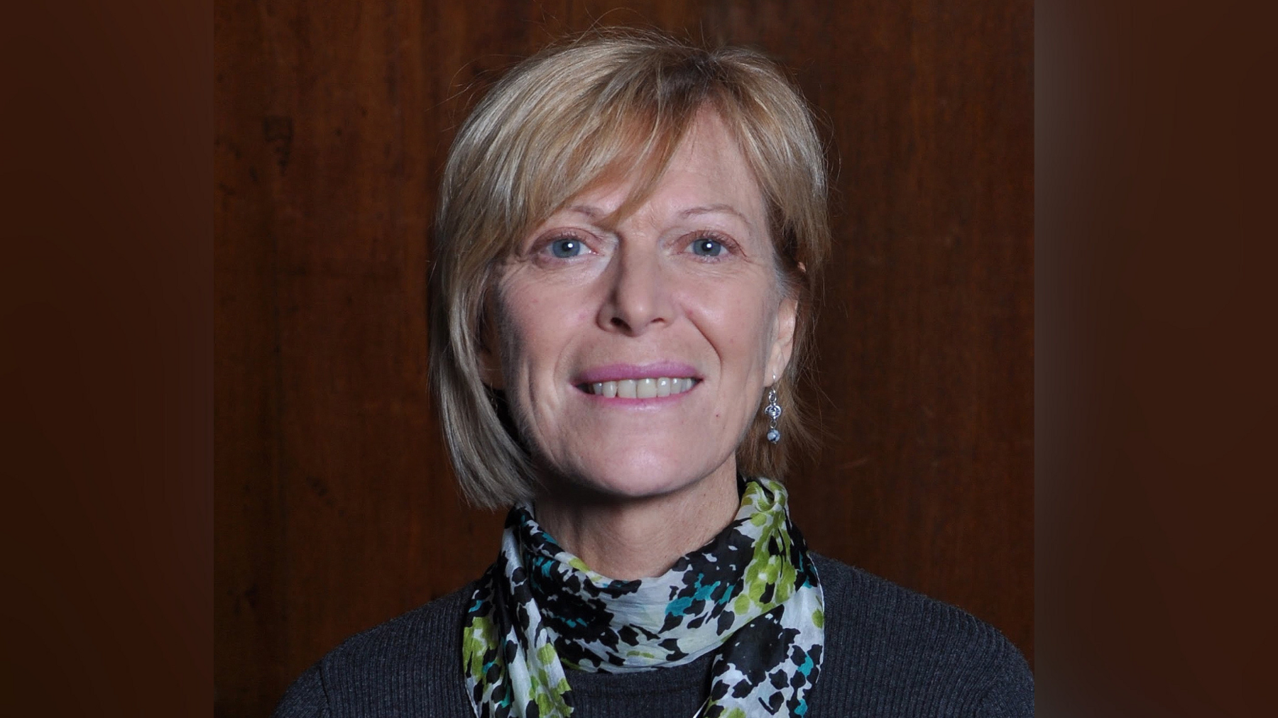 A woman with sandy coloured hair smiling into the camera. She is wearing a blue jumper and a brightly coloured scarf, and the background looks like wood panelling