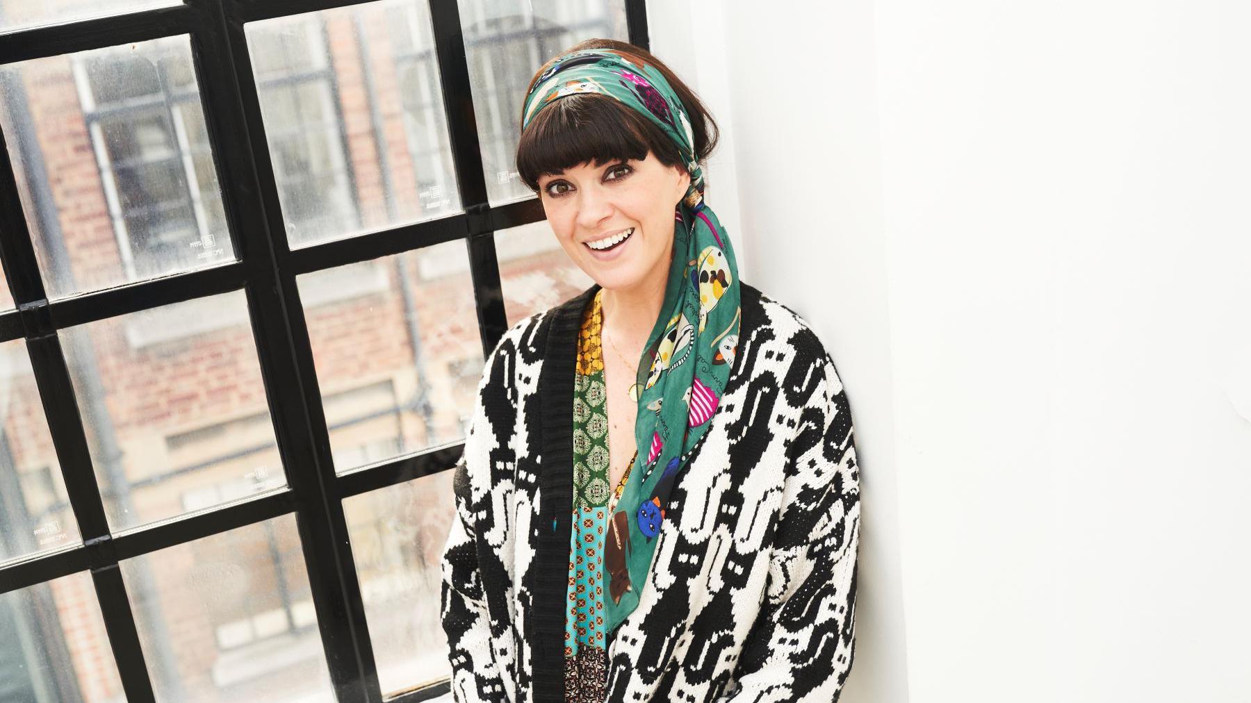 A smiling woman, Dawn O'Porter, with dark hair, heavy eye makeup, and a fringe leans against cast-iron window with a brick building in the background. 

She has a teal headscarf with yellow and purple patterns, and wears a black and white oversized cardigan with a tessellated pattern of cats on it.