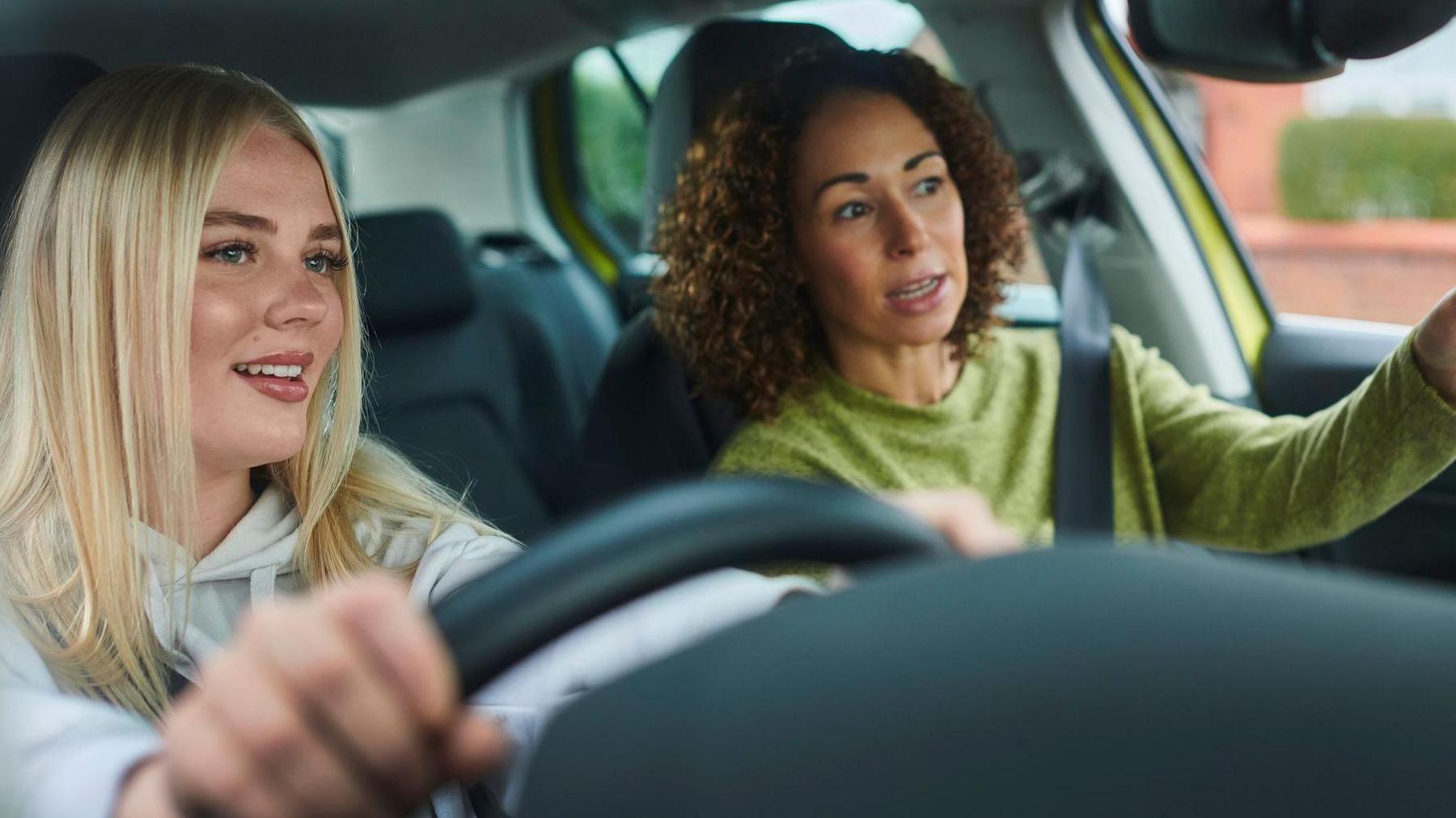 A young woman with blonde hair is behind the wheel of a car. Beside her, a brown-haired woman indicates with her hand as she speaks. 