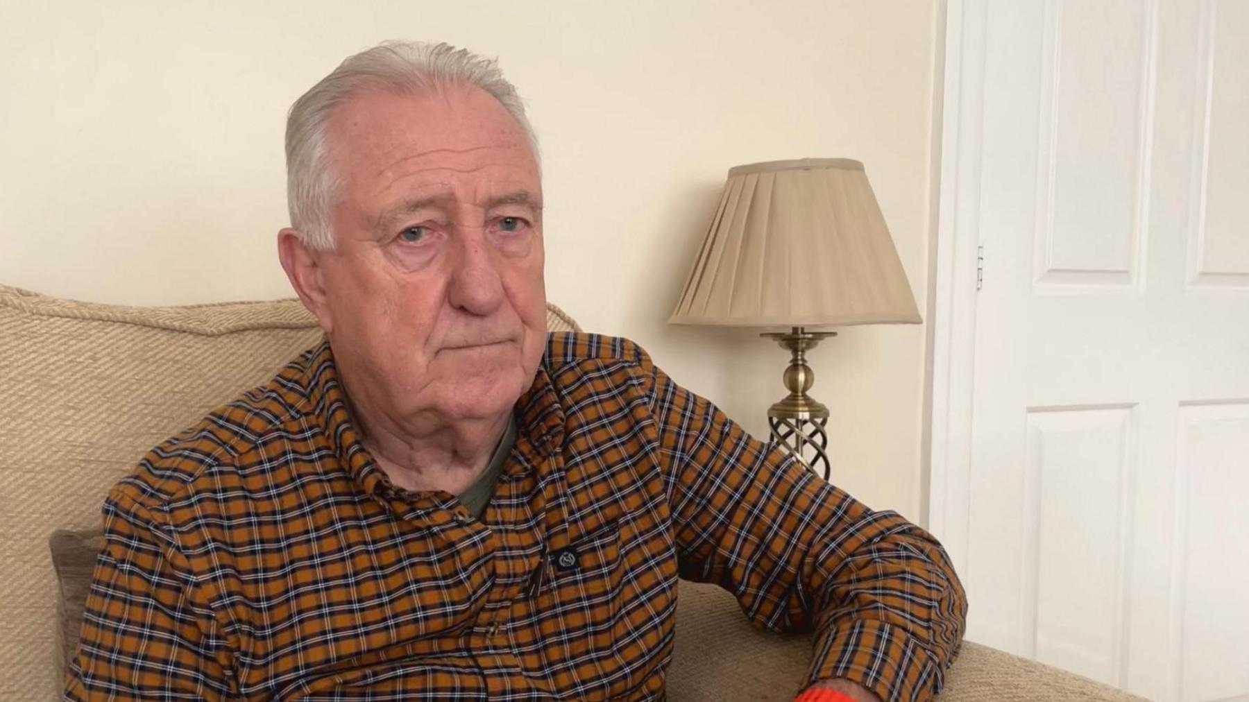 Stuart Porthouse sits in a cream arm chair in front of a lamp in his home. He is wearing a brown-and-black checked shirt.